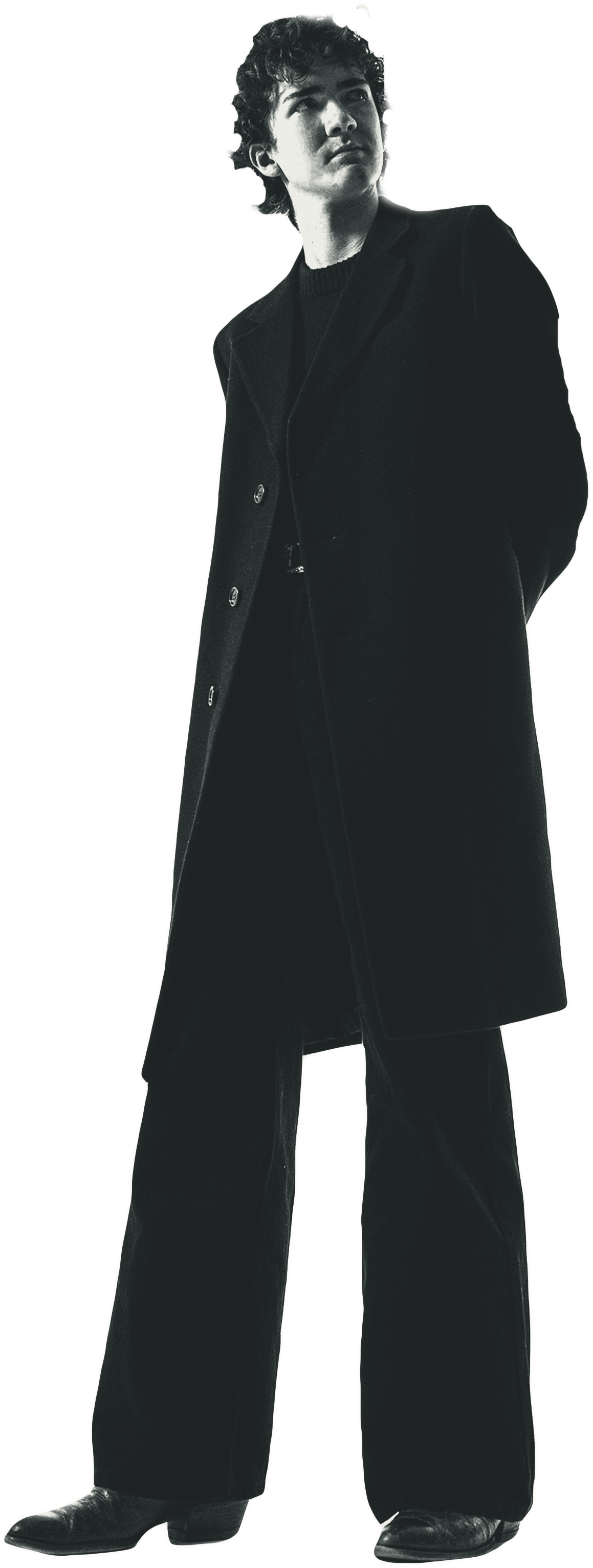 Black-and-white photo of a man with short, curly hair wearing a long black coat, black pants, and black shoes, looking to his left.