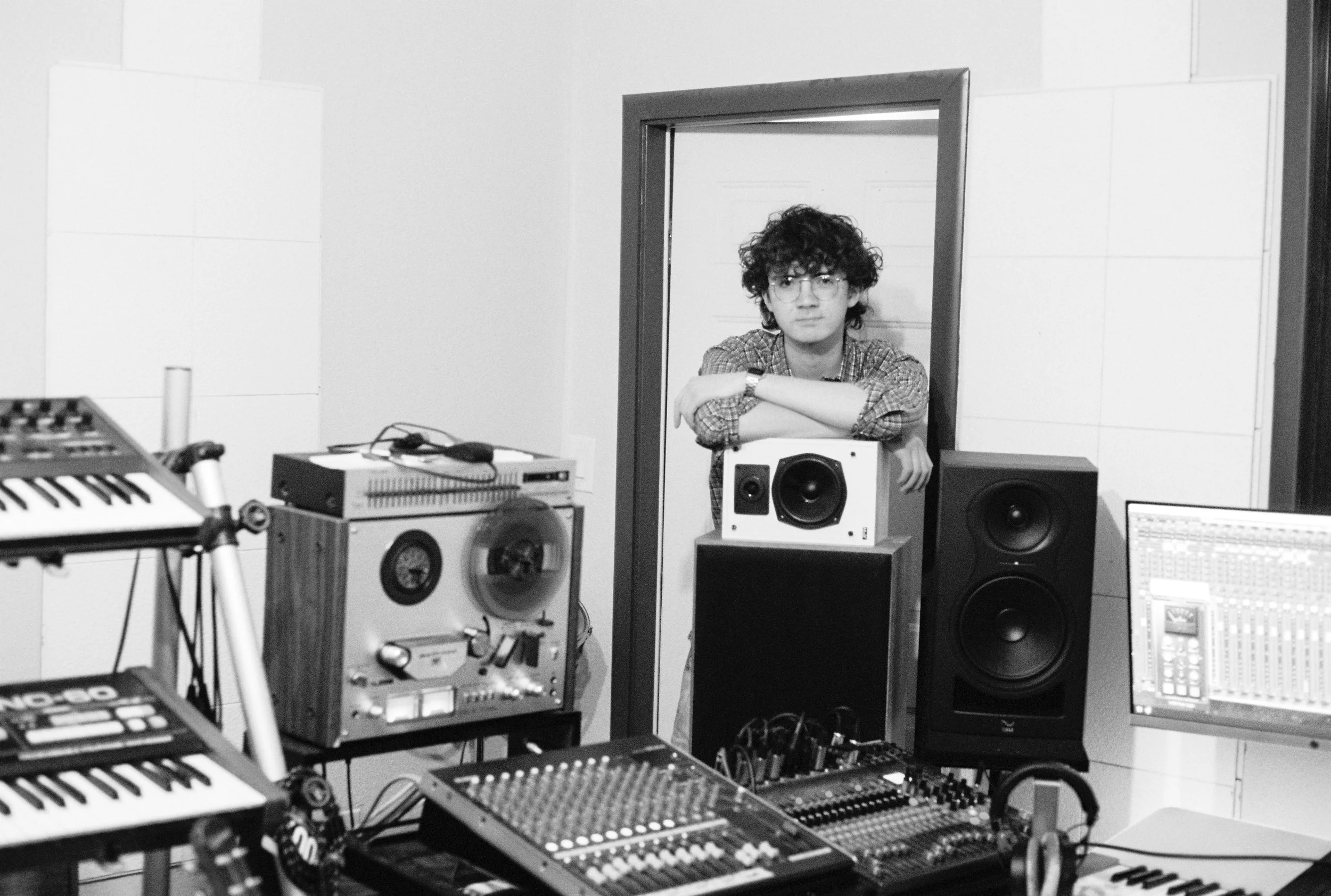 A young man with curly hair and glasses leaning on a piece of studio equipment in a music studio with various electronic music gear, speakers, and a mixing console.
