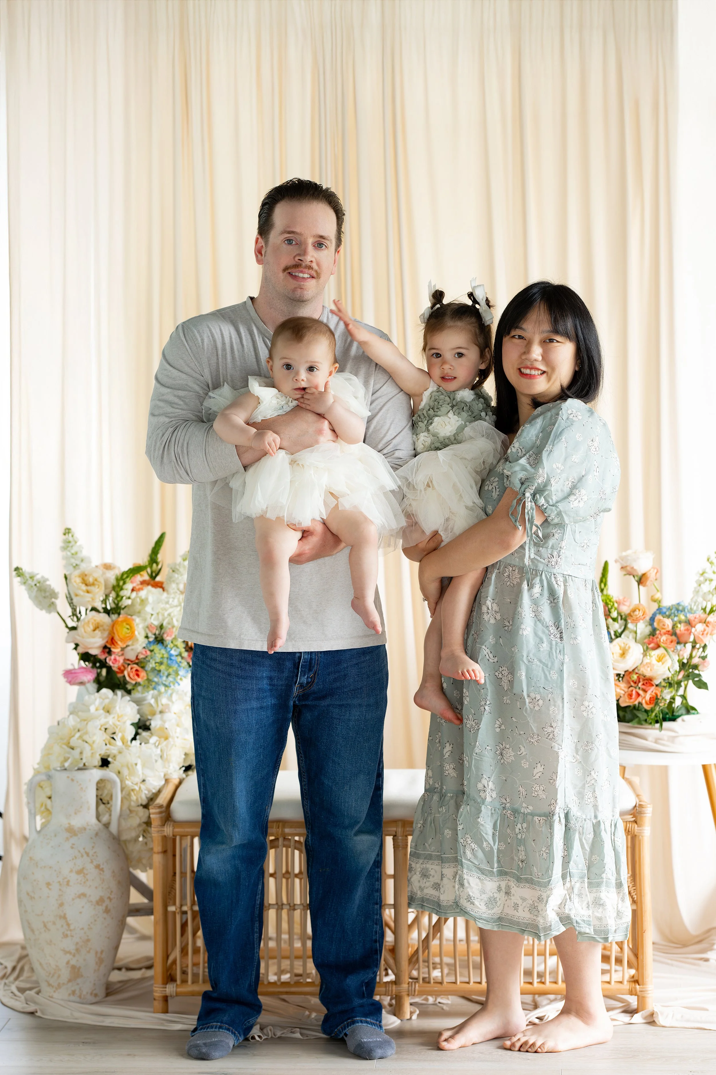 A family of four standing in front of a floral backdrop. The father, with dark hair and a mustache, holds a baby girl dressed in white. The mother, with shoulder-length dark hair, is holding a young girl with pigtails and bows. The family is smiling and dressed in light-colored, floral, and pastel clothing.