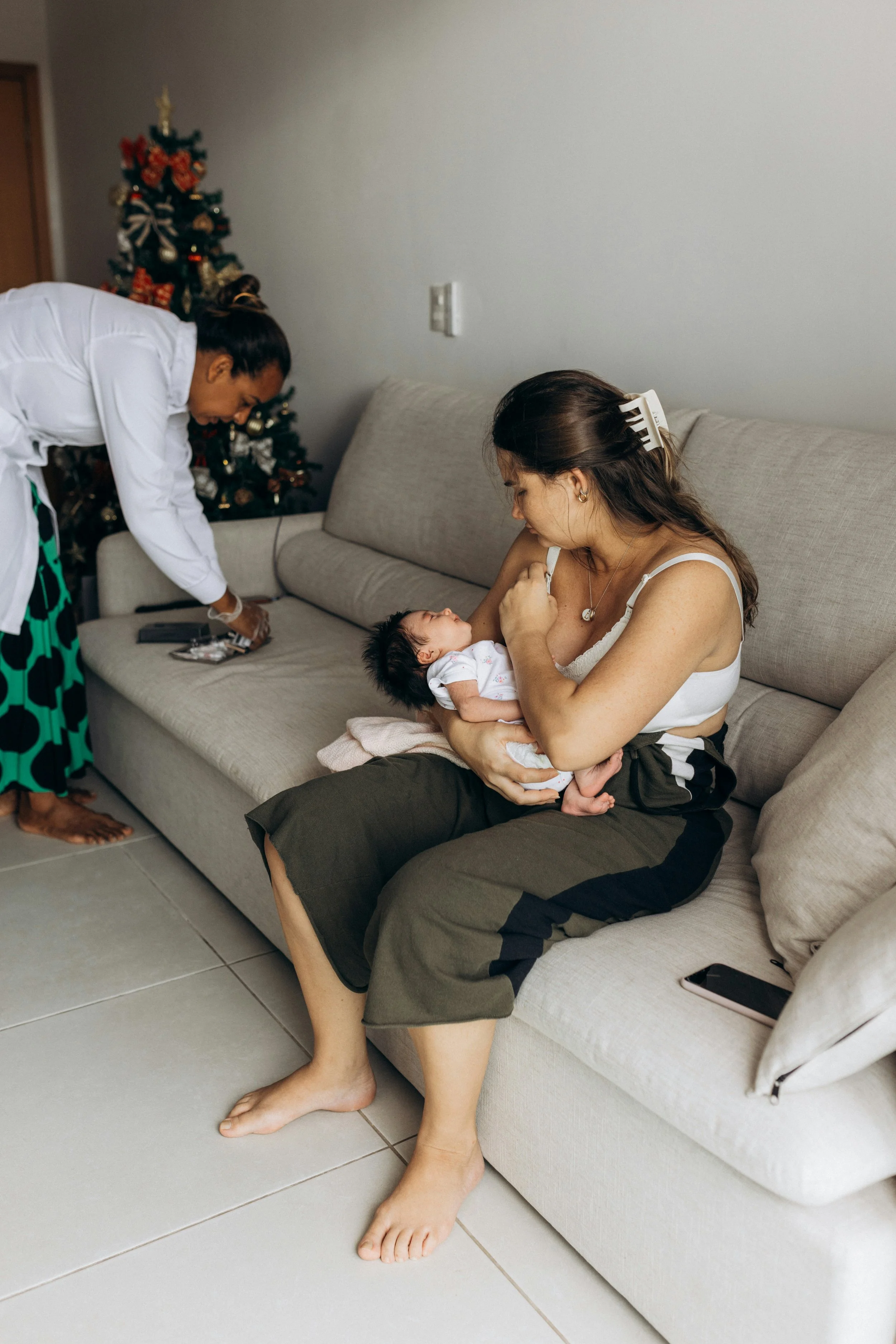 A woman sitting on a couch holding a baby, with hair clips in her hair, and a Christmas tree in the background. A person in a white shirt is near the couch, and the room has tiled flooring.