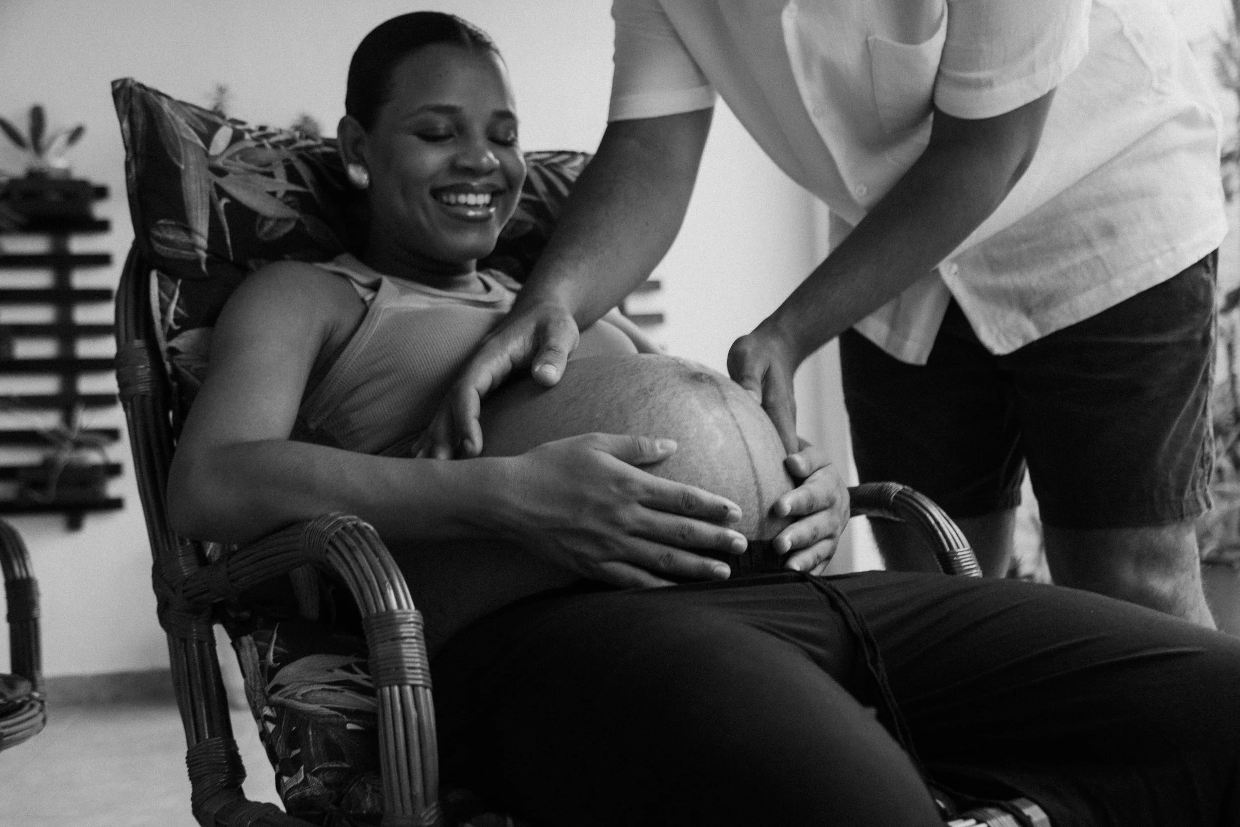 A woman with short hair smiling and sitting in a chair, receiving a pregnancy check-up from a male healthcare provider, with hands on her pregnant belly. The setting appears to be a medical or home environment.
