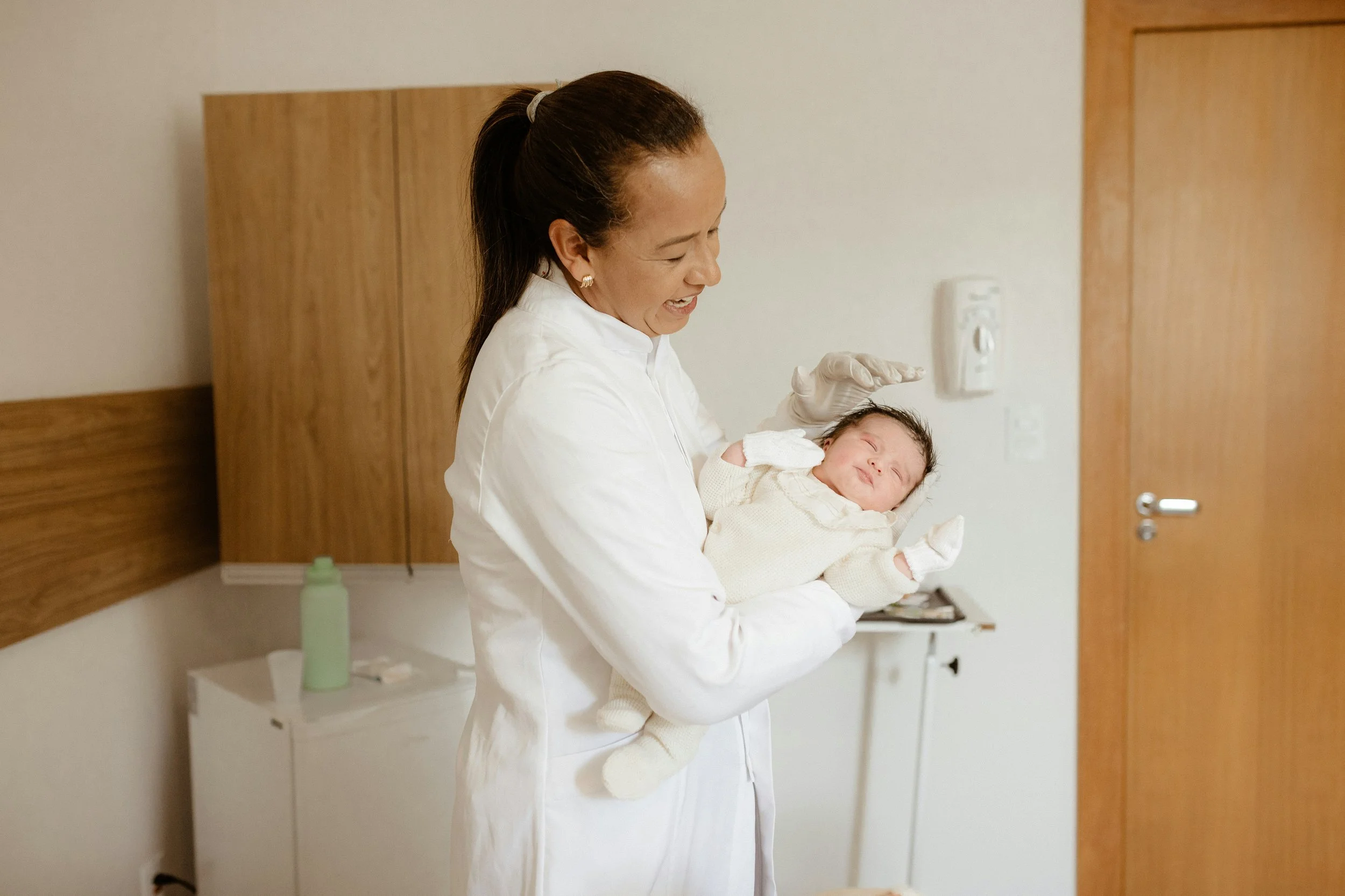 A woman wearing a white medical coat and gloves holding a newborn baby in a hospital room.