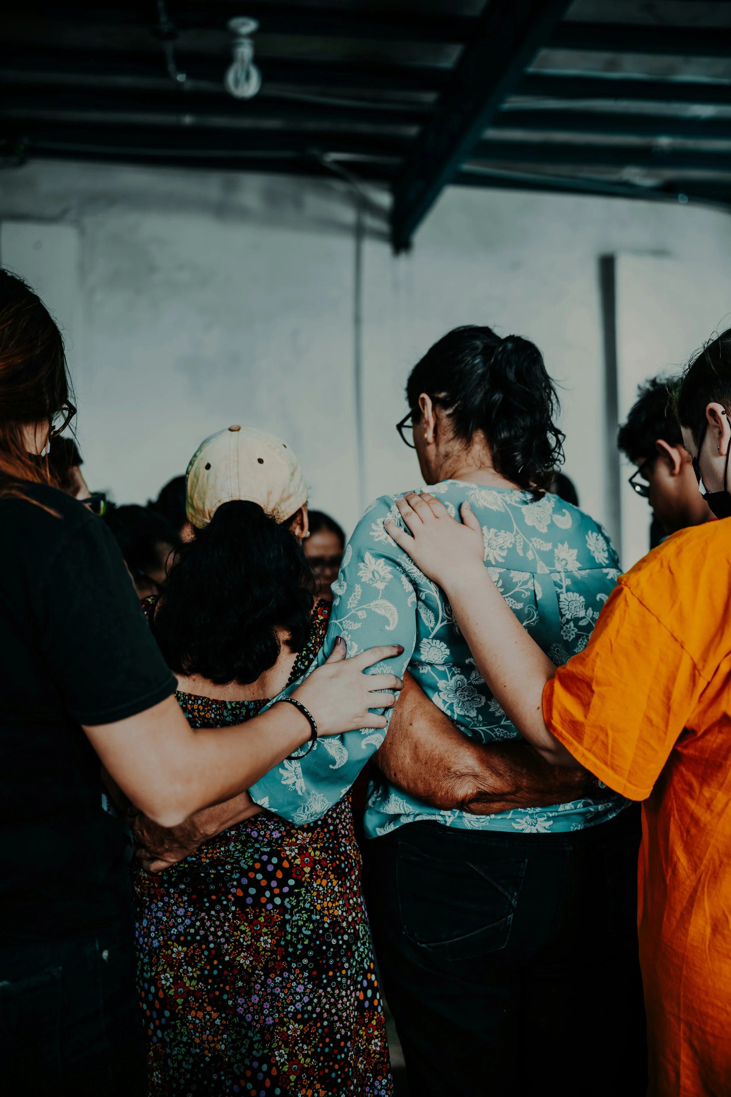 A group of people gathered closely together during a moment of prayer or support, with one person comforting and praying for an elderly woman.