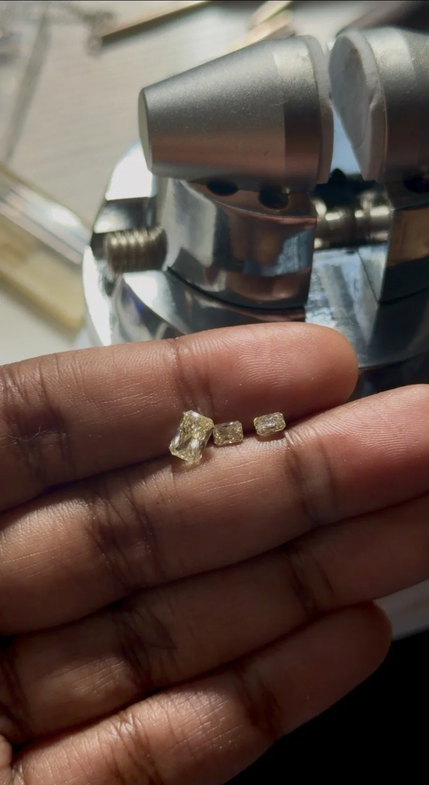 Person's hand holding small, yellow radiant cushion cut diamonds near a metallic machine or tool.