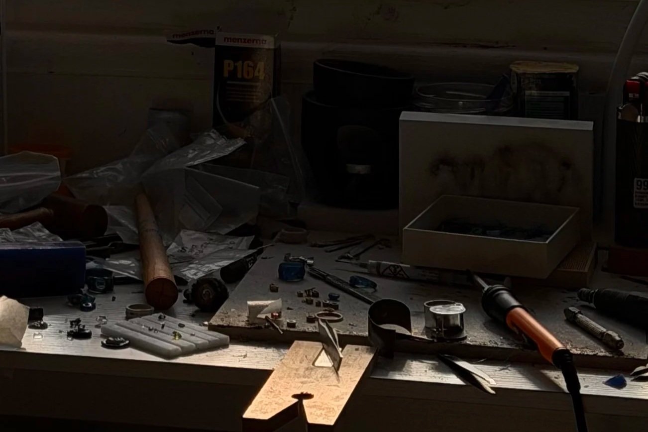 Disorganized jewellery workbench with tools, electronics, and clutter, illuminated by a focused light.