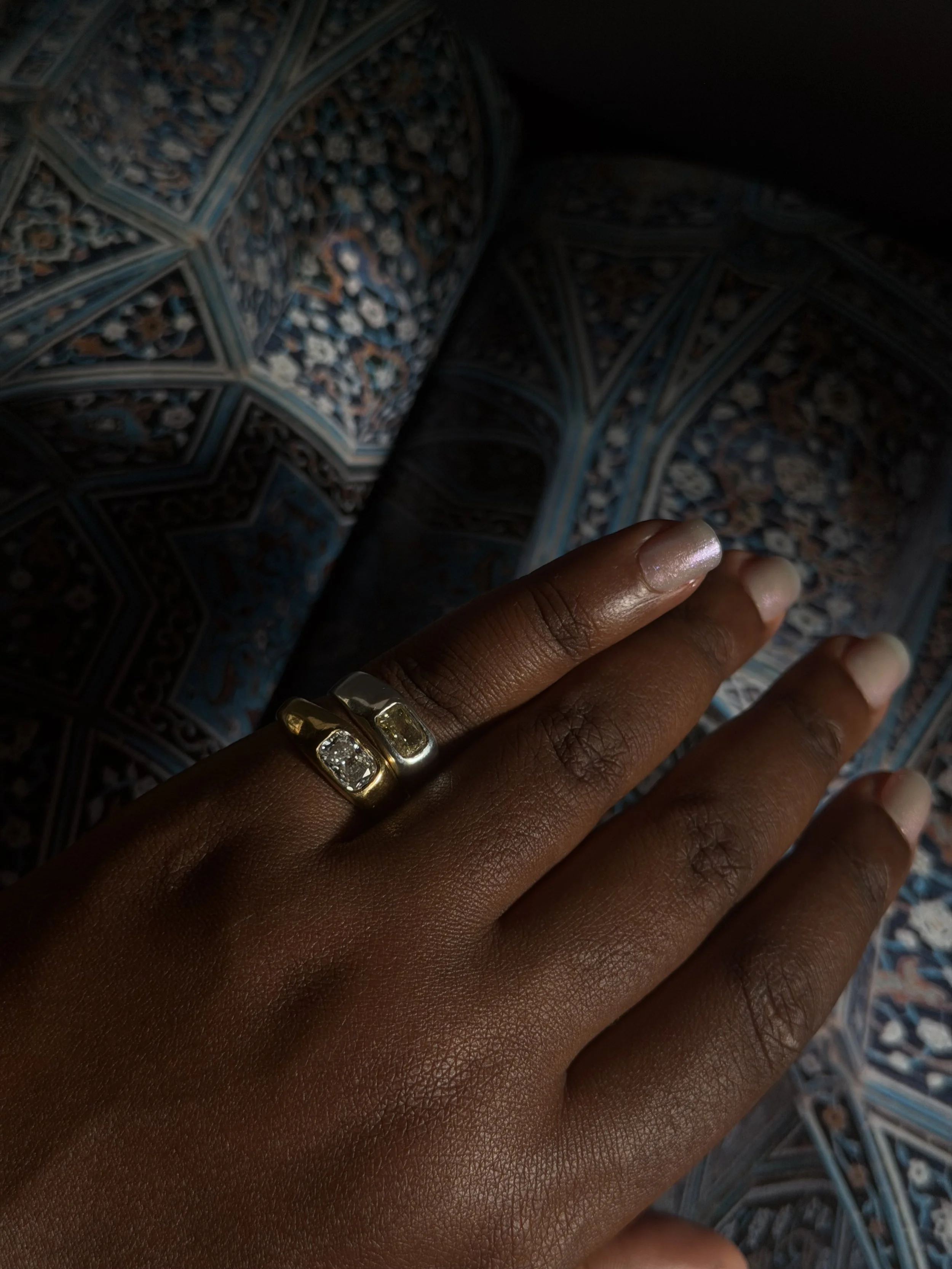 Hand with dark skin wearing a gold ring with three large clear stones, resting on a patterned fabric with intricate blue, white, and brown designs.