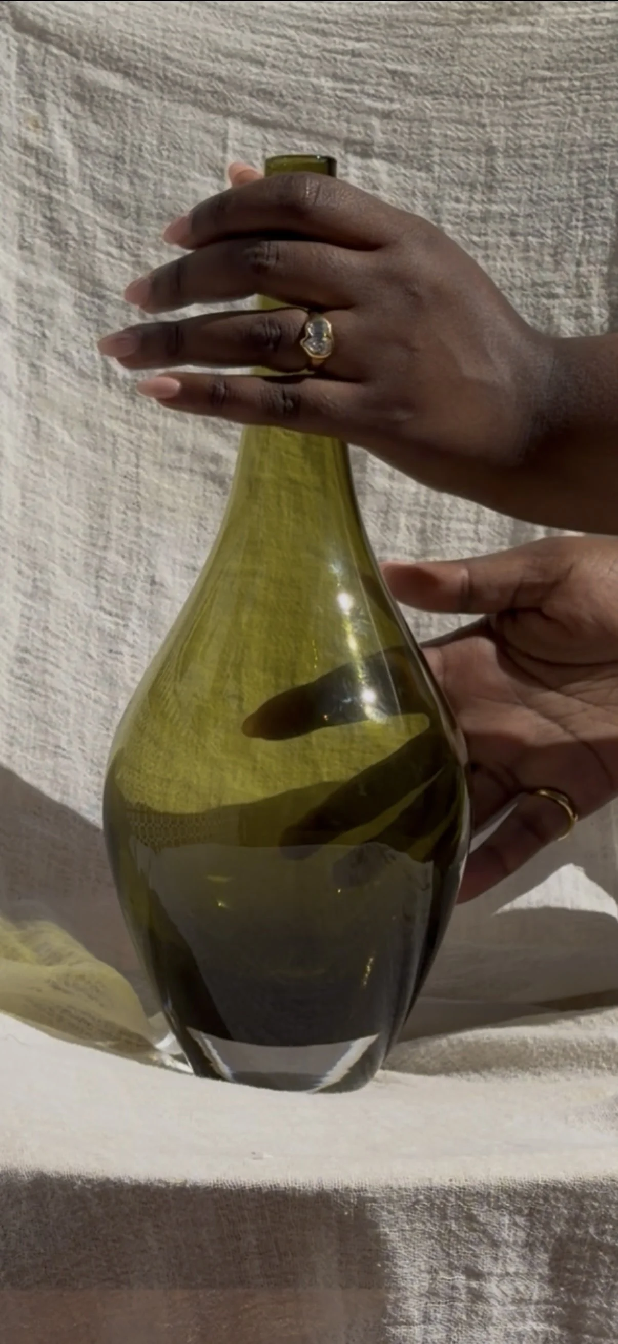 A hand holding a green glass vase against a textured white wall. Wearing an organic gold diamond engagement ring.