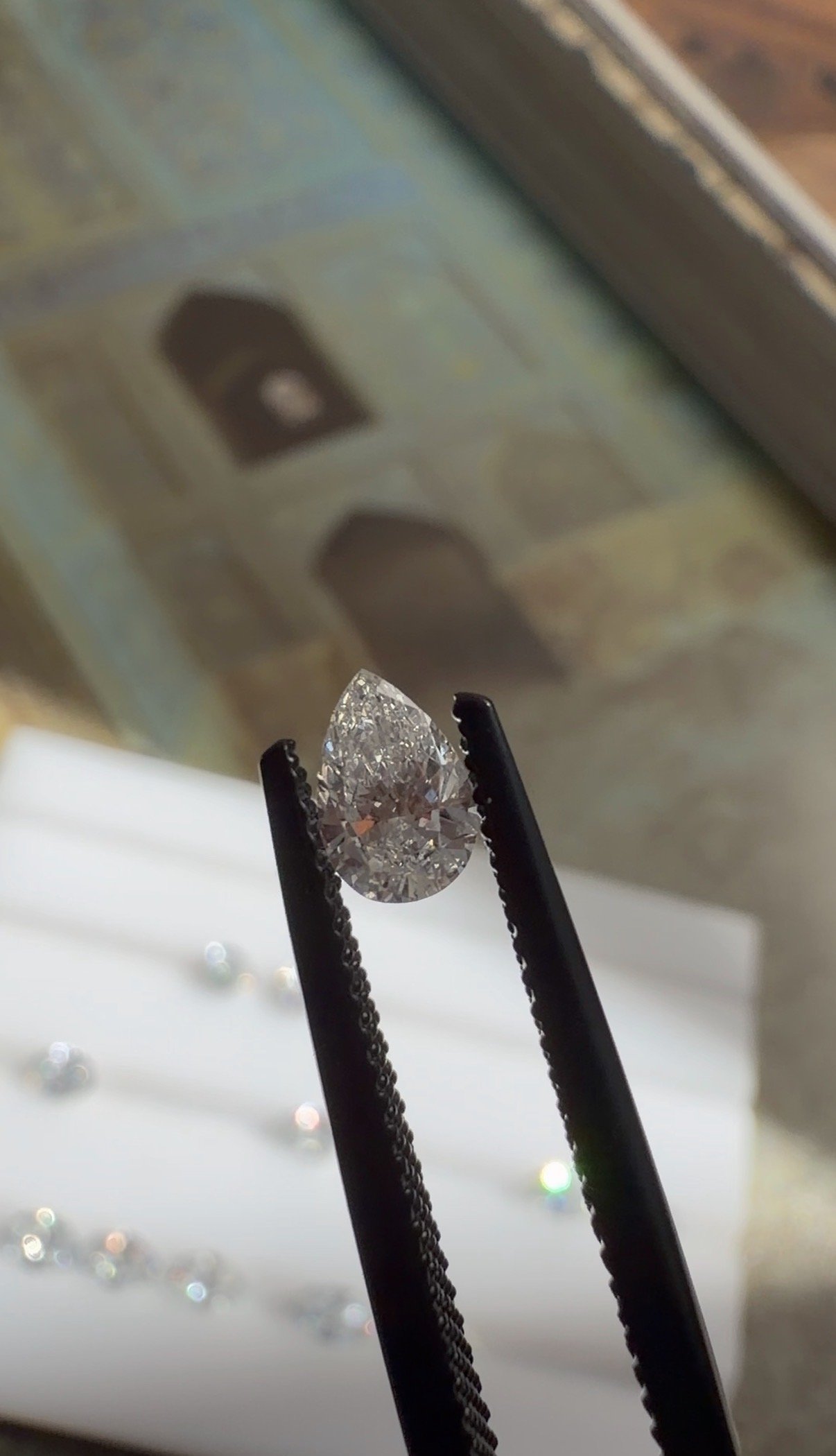 Close-up of a pear-shaped diamond being held with black tweezers, with blurred background including a window and some scattered smaller gemstones or reflections.