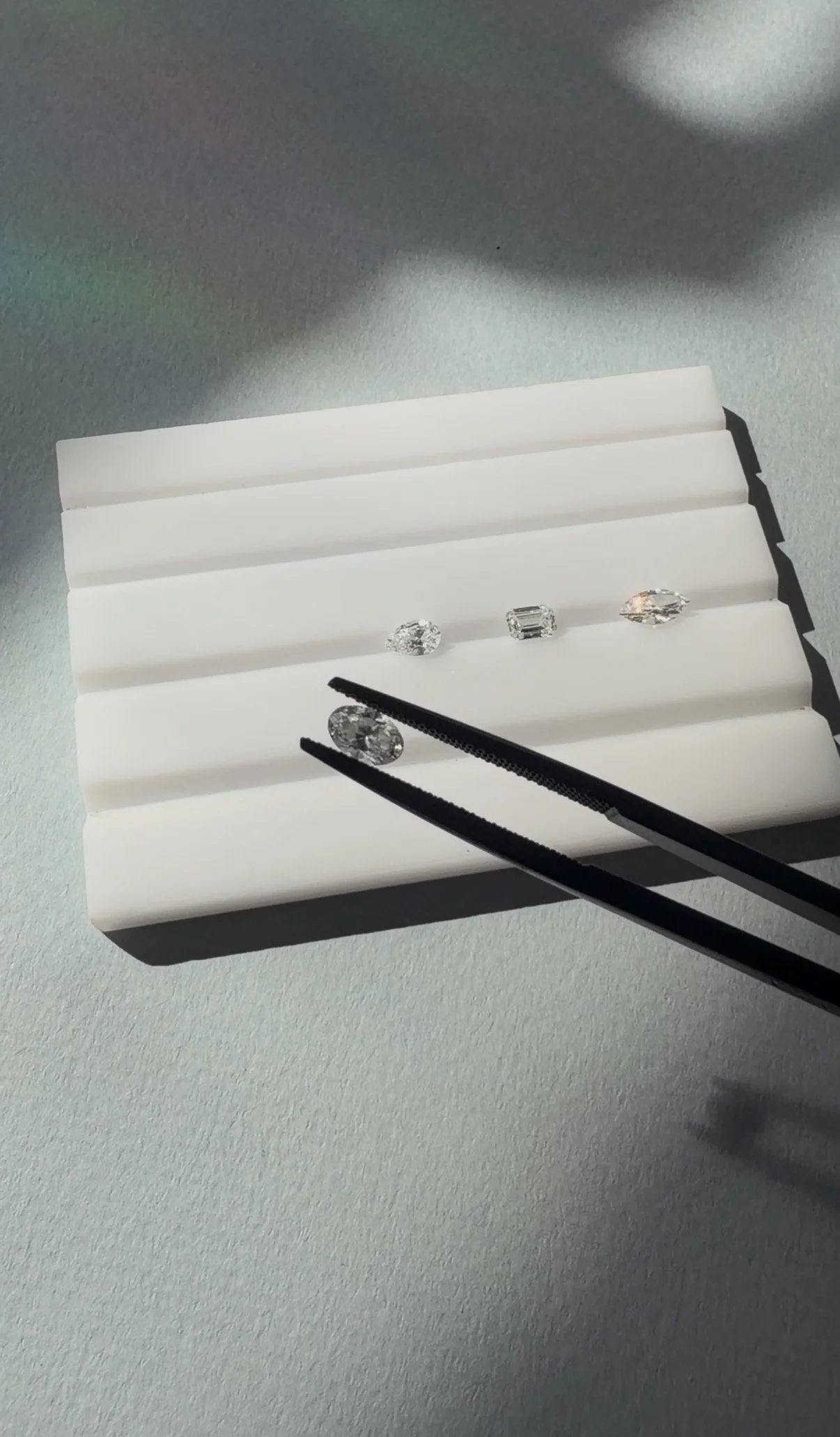 Three diamonds on a white display tray with black tongs holding one diamond, illuminated by light on a plain background.