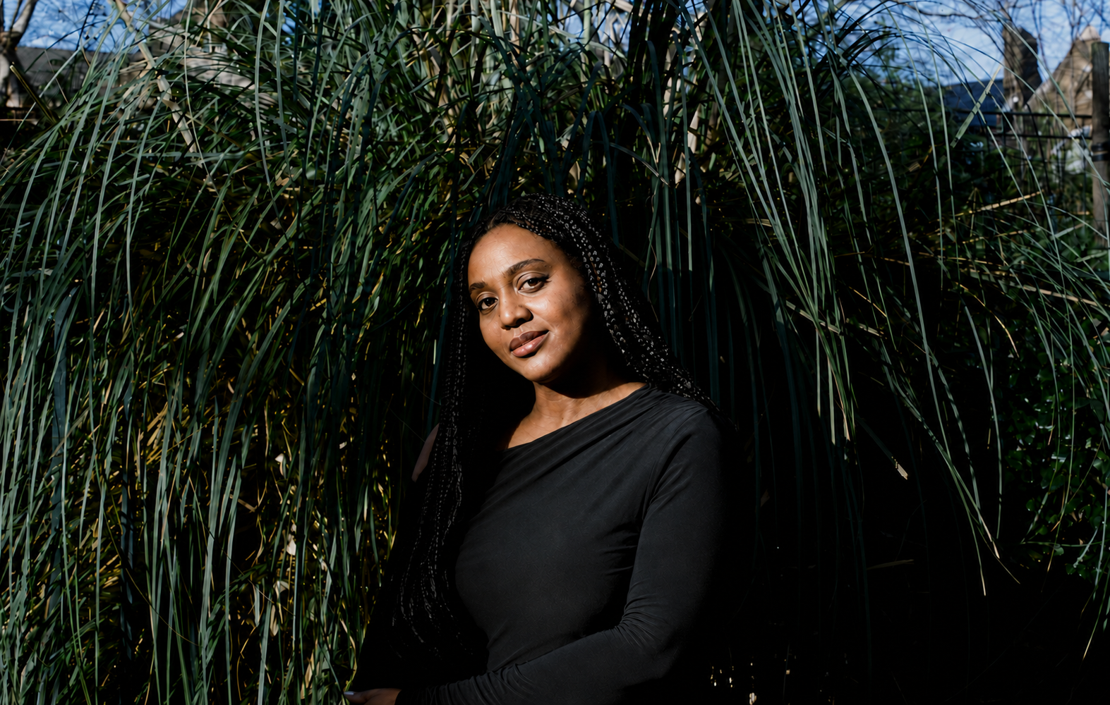 A woman with long braided hair, wearing a black top, stands against a background of tall green grass and plants, outdoors in daylight.