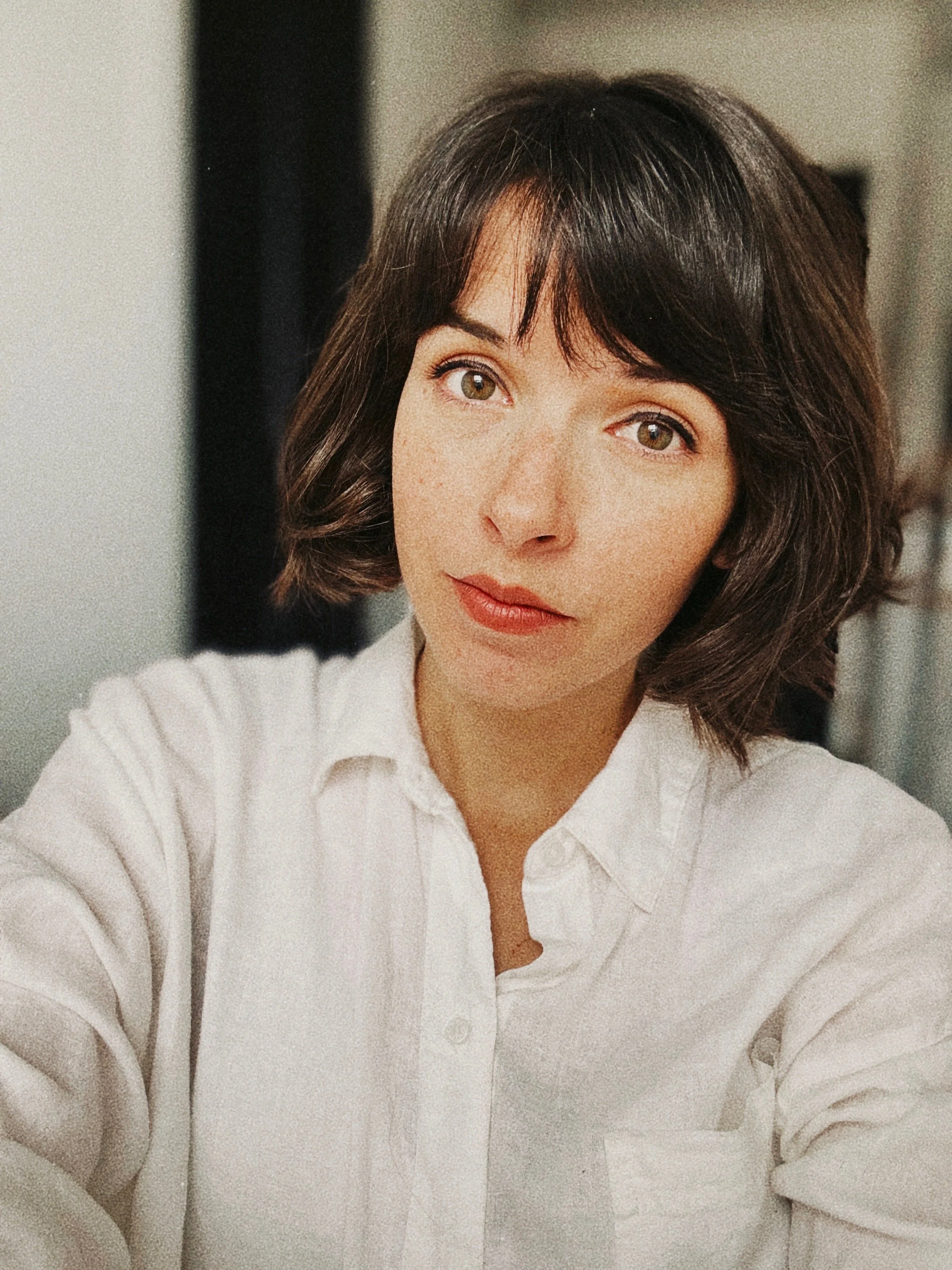 A woman with dark brown, wavy hair and hazel eyes wearing a white button-up shirt, looking at the camera.