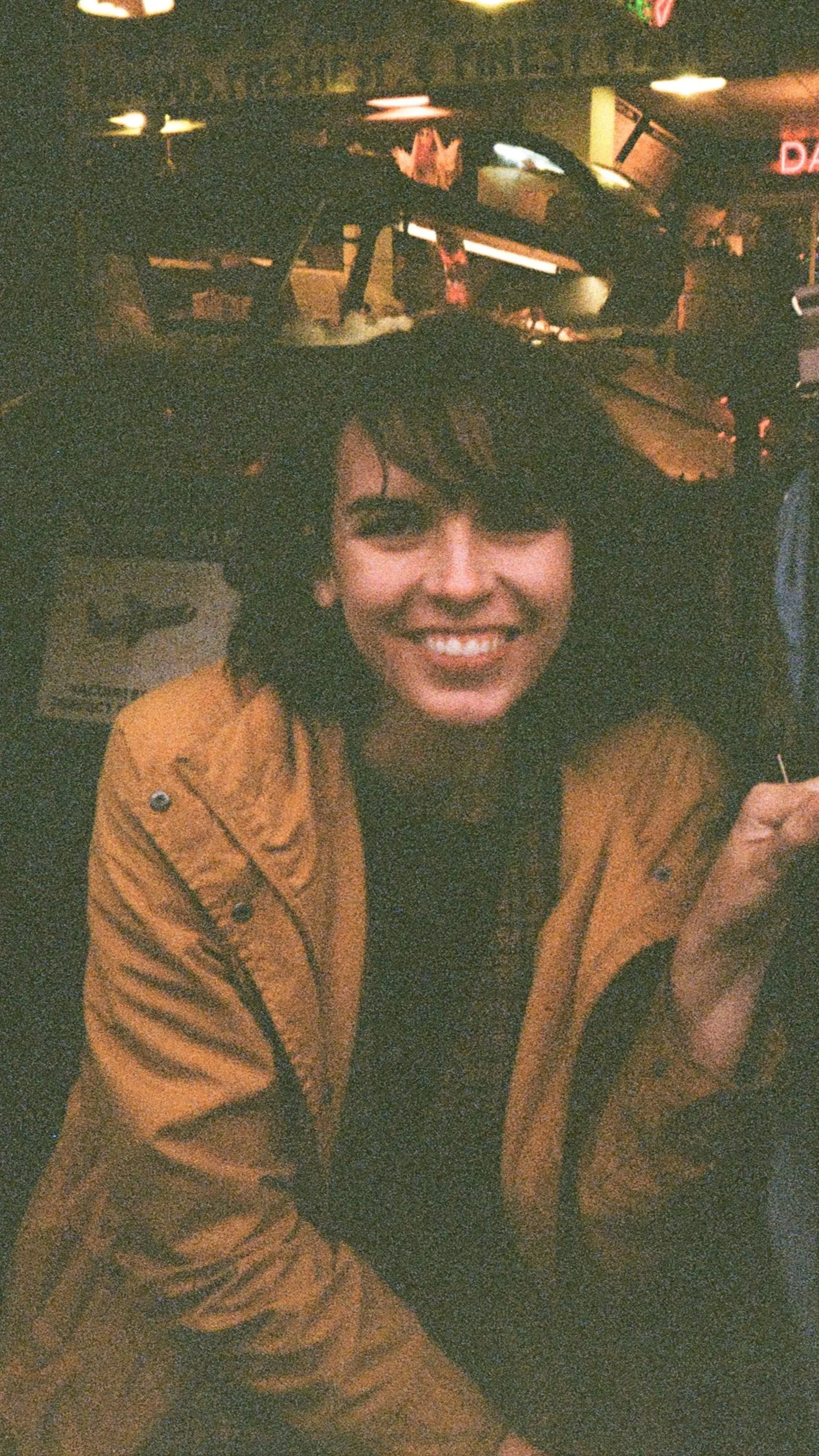 A woman with short dark hair and a bright smile, wearing a tan jacket, sitting in a dimly lit restaurant or bar.