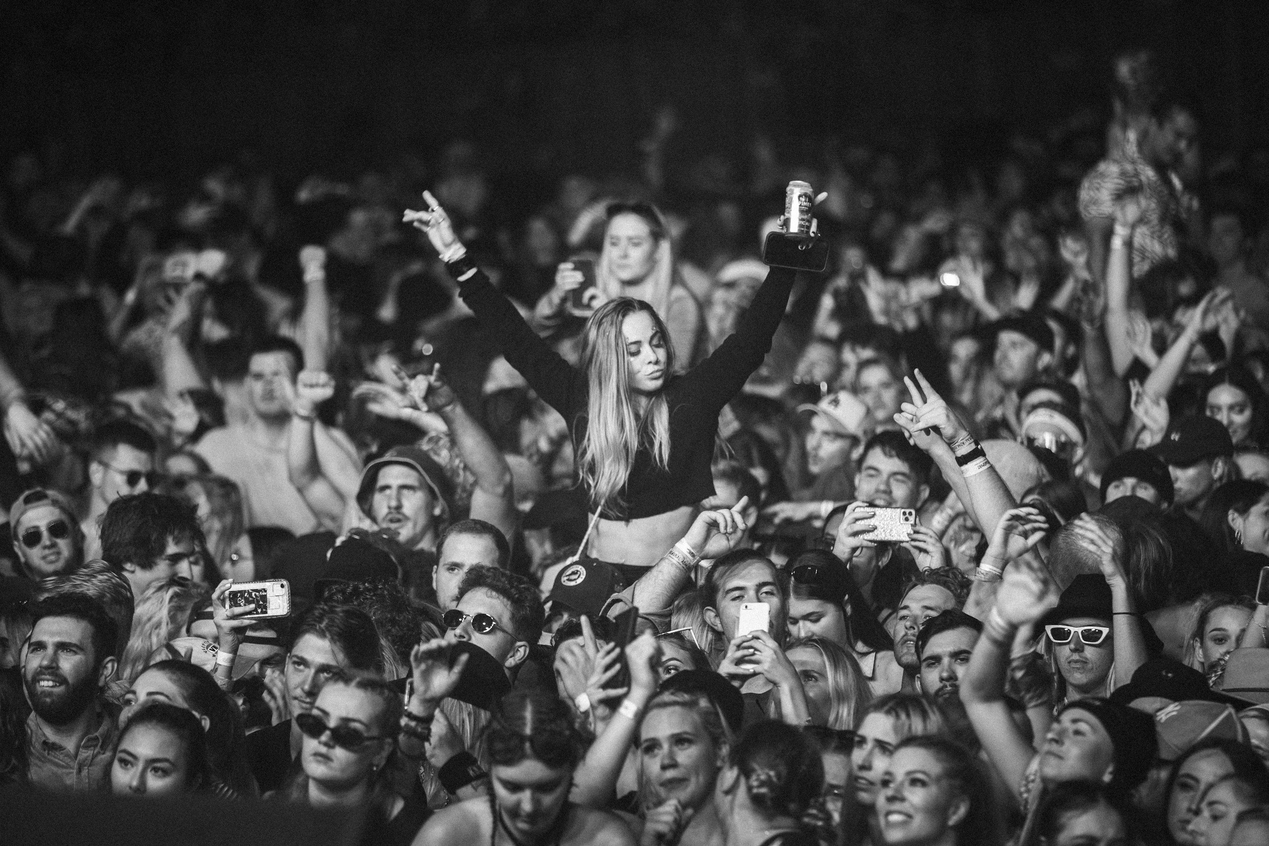 A black and white photo of a crowded concert or music festival. A young woman in the center holds a drink and raises her hand in a peace sign, appearing to enjoy the event. Many people around her are taking photos or simply watching the performance, with some wearing sunglasses or headphones.