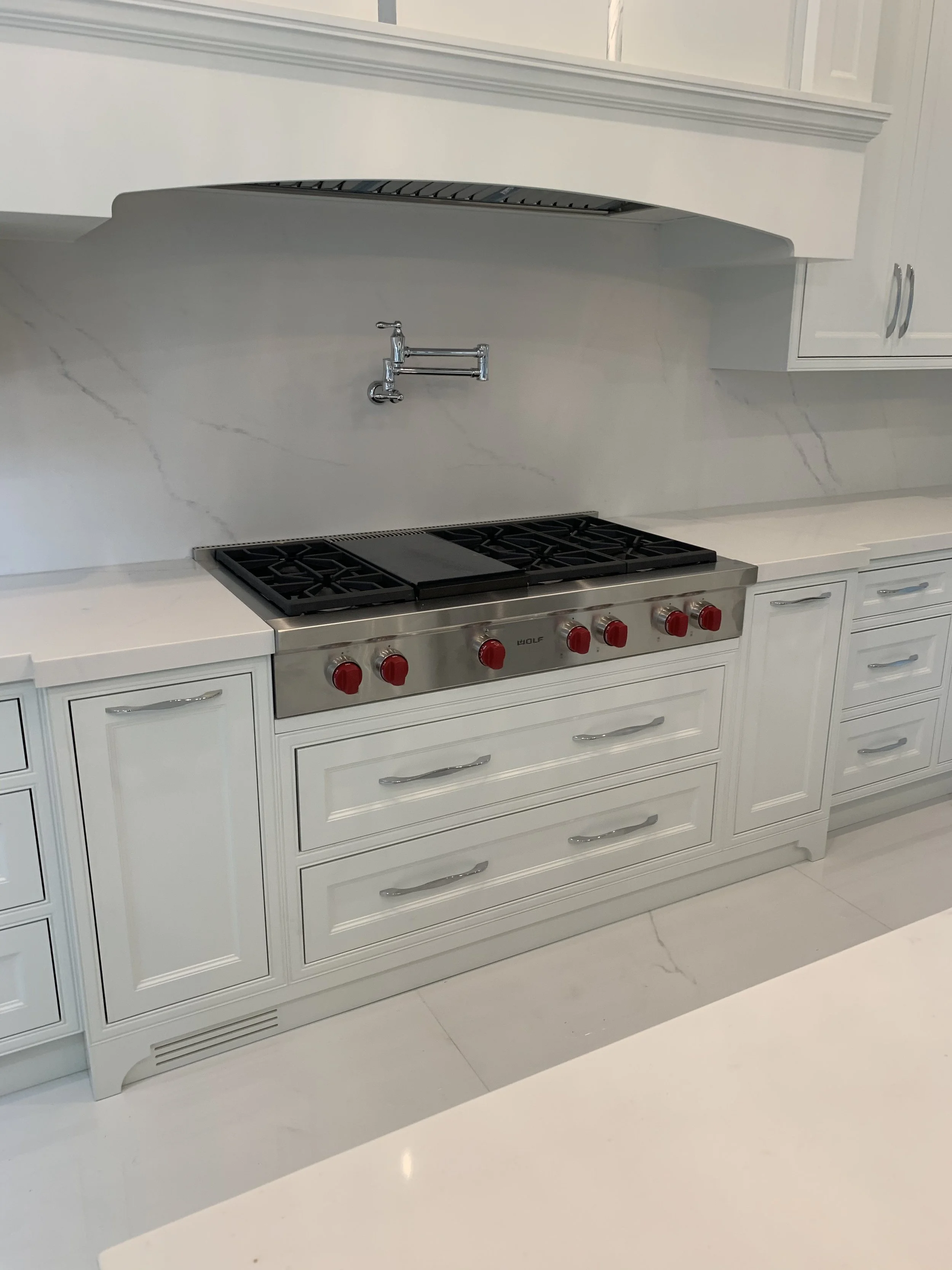 A kitchen with white cabinets, a countertop, a stainless steel stove with red knobs, and a marble backsplash, with floor and countertop in white.