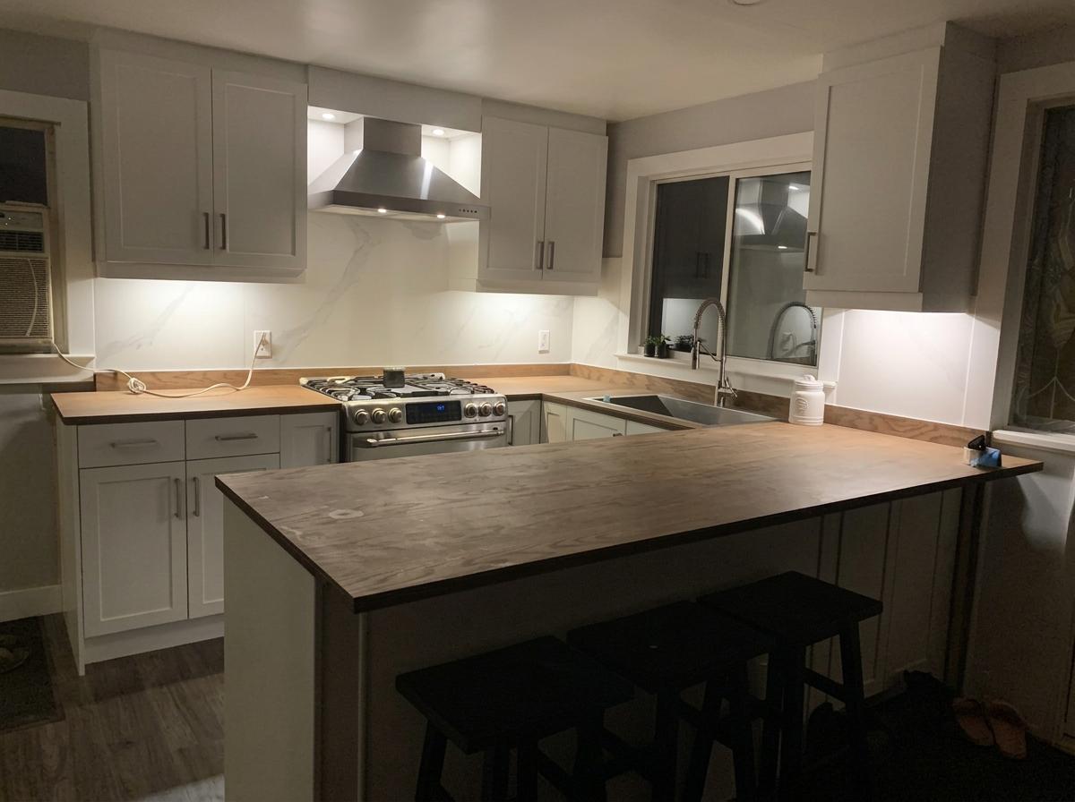 Modern kitchen with white cabinets, marble backsplash, a wooden countertop, stainless steel stove and range hood, a double window above the sink, and black stools at a kitchen island.