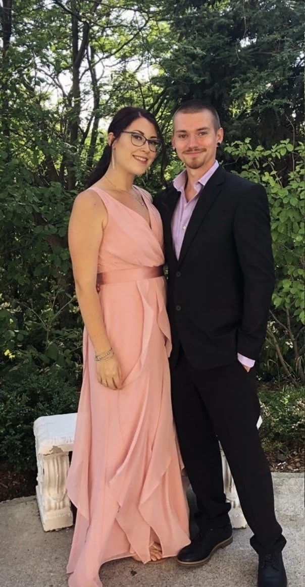 A woman in a pink dress standing next to a man in a black suit, outdoors with greenery in the background.
