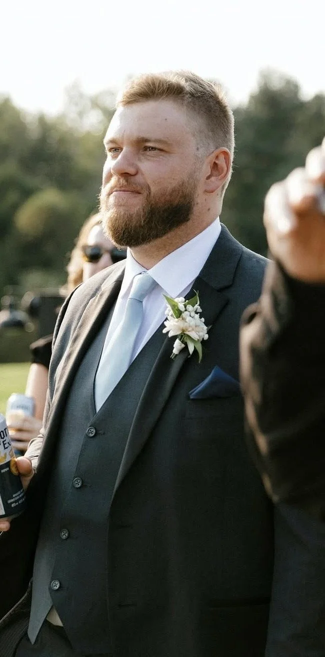 A man with a beard and short hair wearing a dark suit, light blue tie, and a white dress shirt, standing outdoors during daytime, with a floral boutonniere on his lapel and a pocket square in his suit pocket.