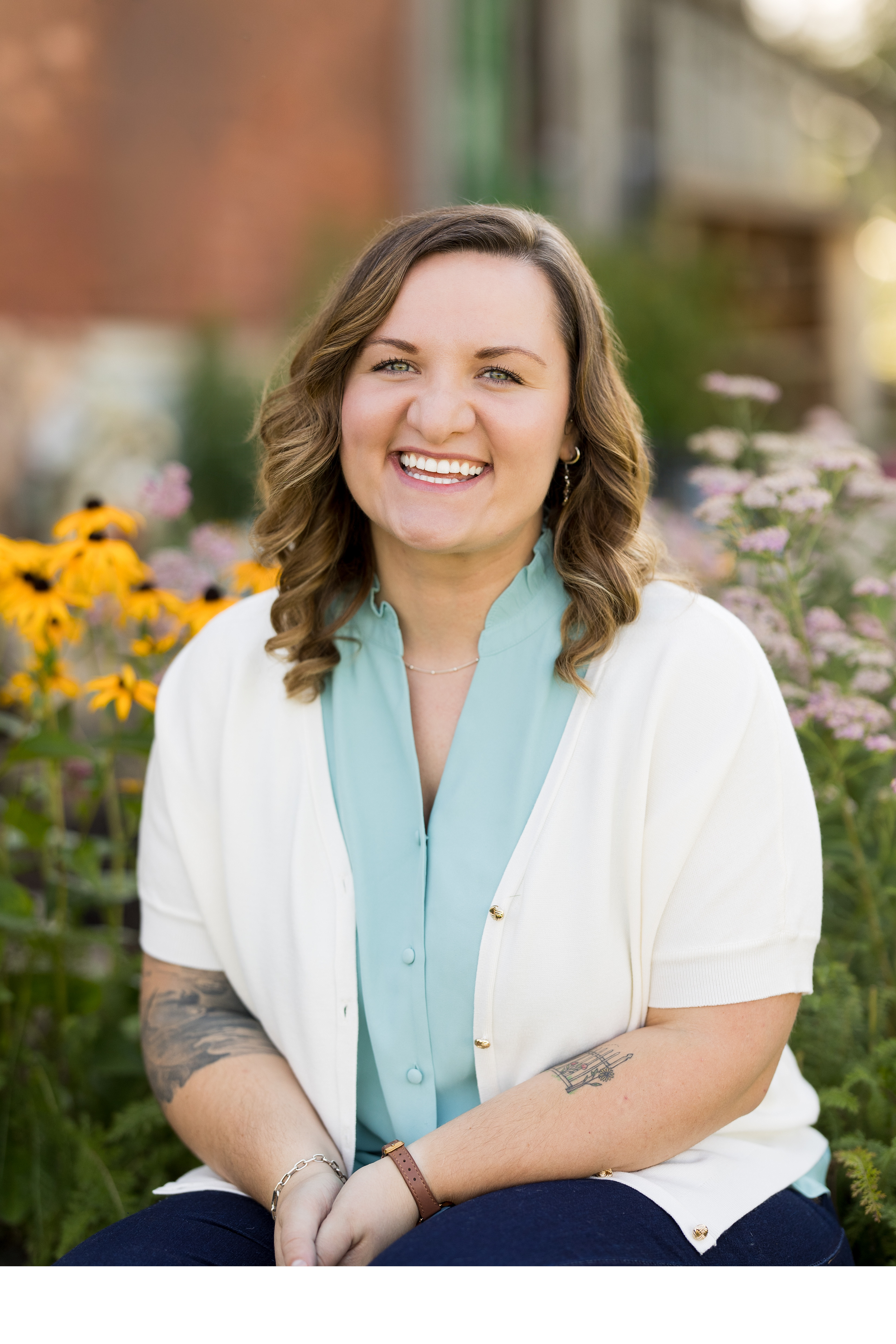 A young woman with shoulder-length brown hair, smiling, sitting outdoors surrounded by flowers and greenery, wearing a white cardigan over a light blue blouse, with tattoos on her arms, and accessories including earrings, a bracelet, and a watch.