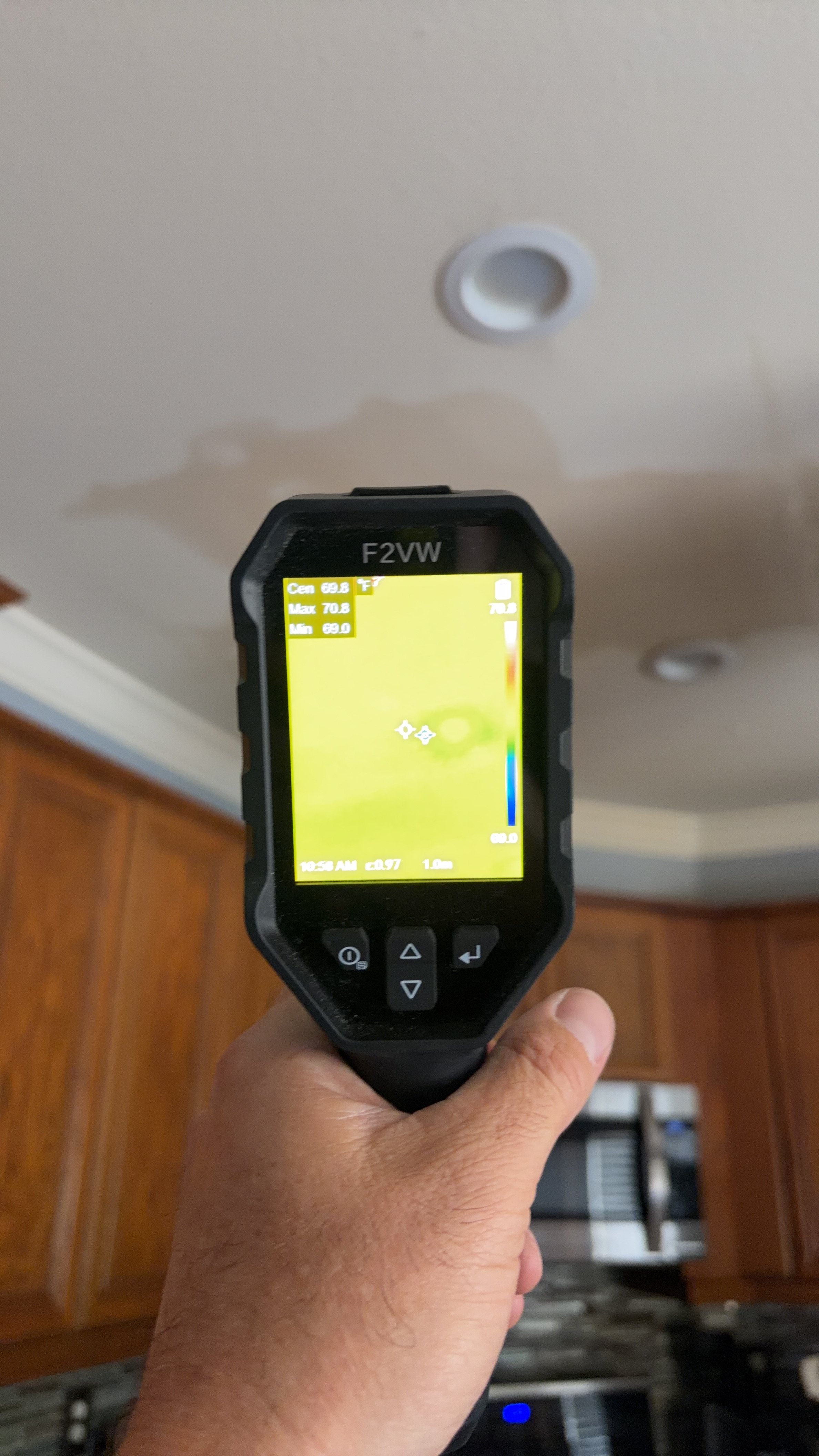 A handheld thermal imaging camera showing a color temperature map of the ceiling, with wooden cabinets in the background.