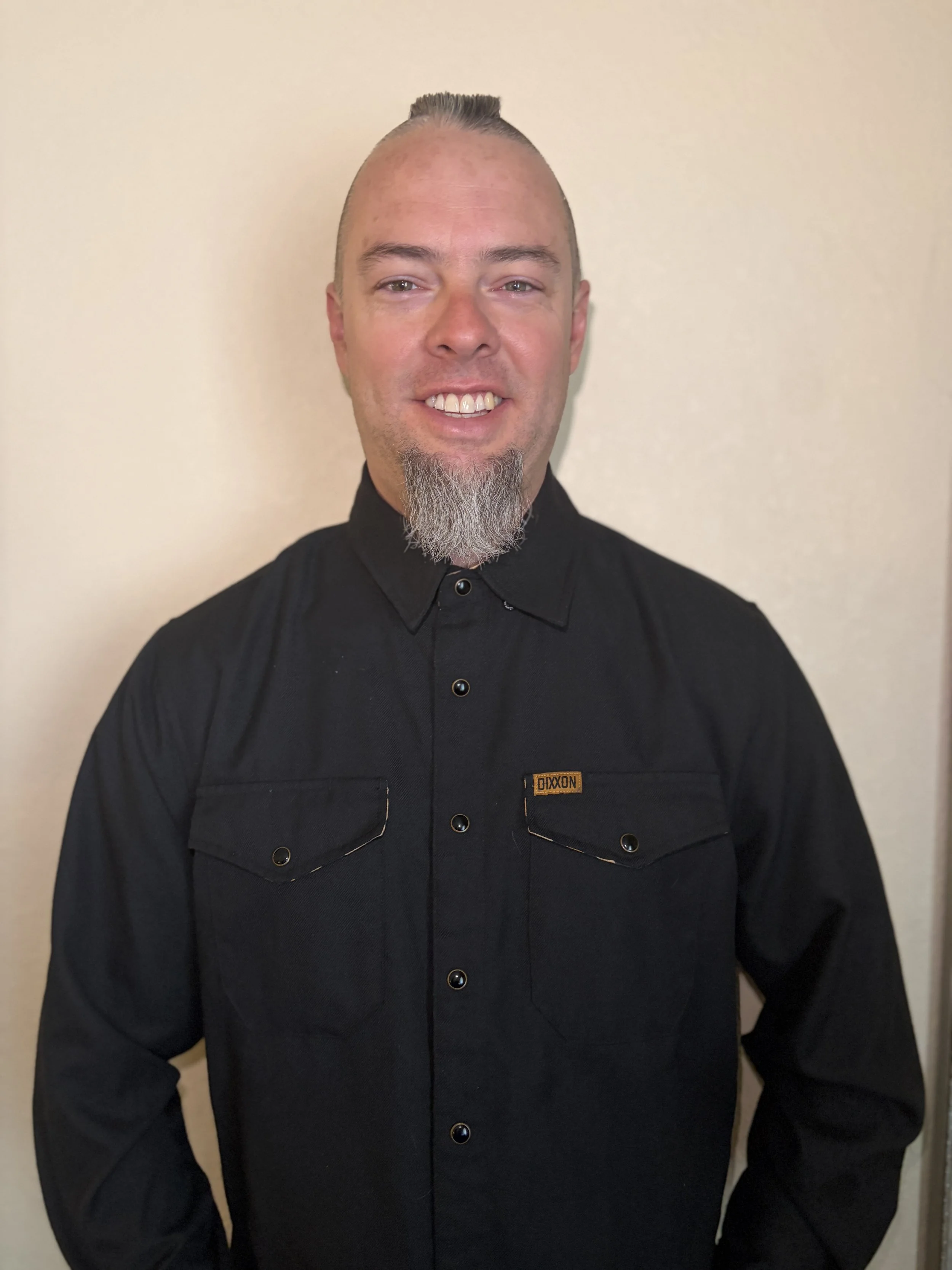 A home inspector/business owner in a black button-up shirt with a friendly smile standing in front of a beige wall