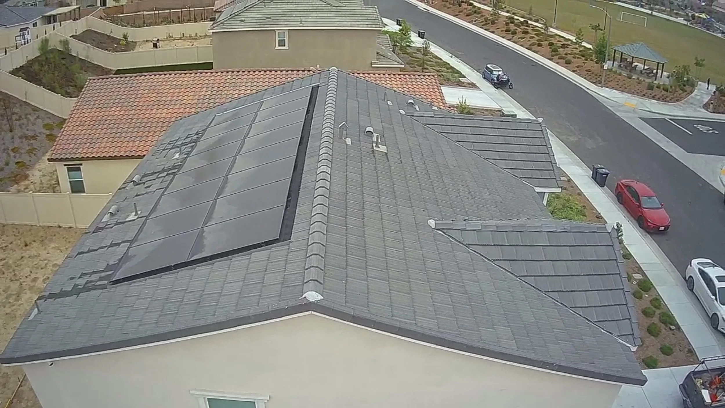 View of rooftops with solar panels on a residential house in a neighborhood with streets, parked cars, a fenced backyard with gardening beds, and a small park with a gazebo.