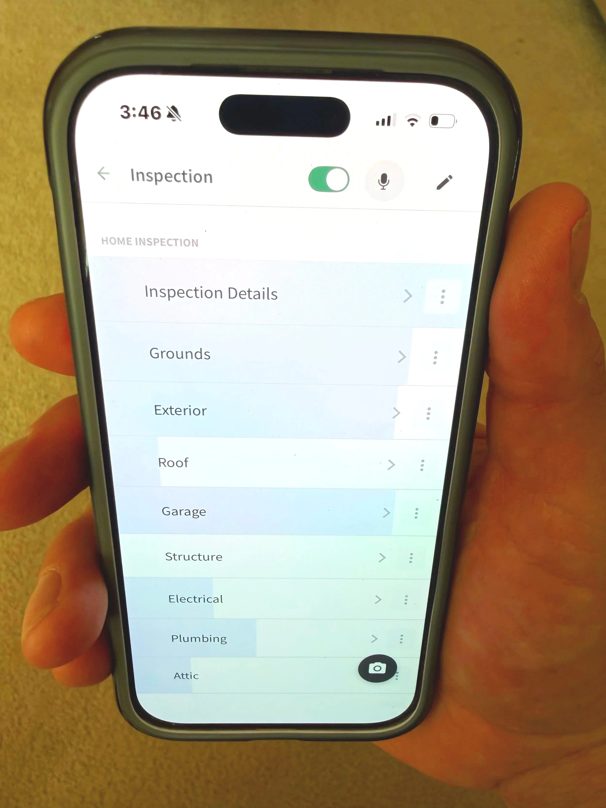 A person holding a smartphone displaying a home inspection application menu with options for various inspection categories such as Grounds, Exterior, Roof, Garage, Structure, Electrical, Plumbing, and Attic.