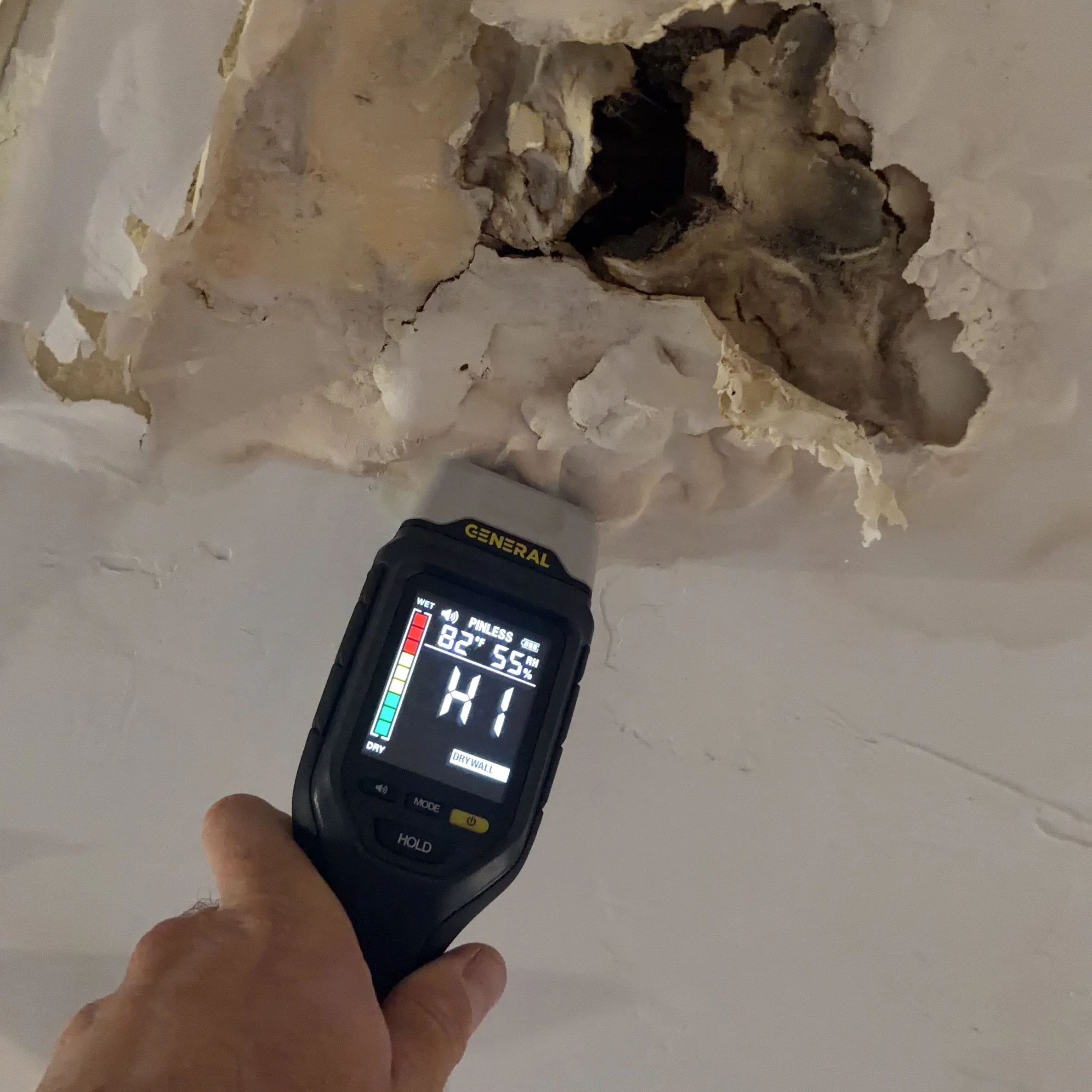 A person's hand holding a digital moisture meter displaying 82% moisture level, aimed at a hole in a wall with exposed drywall and insulation.