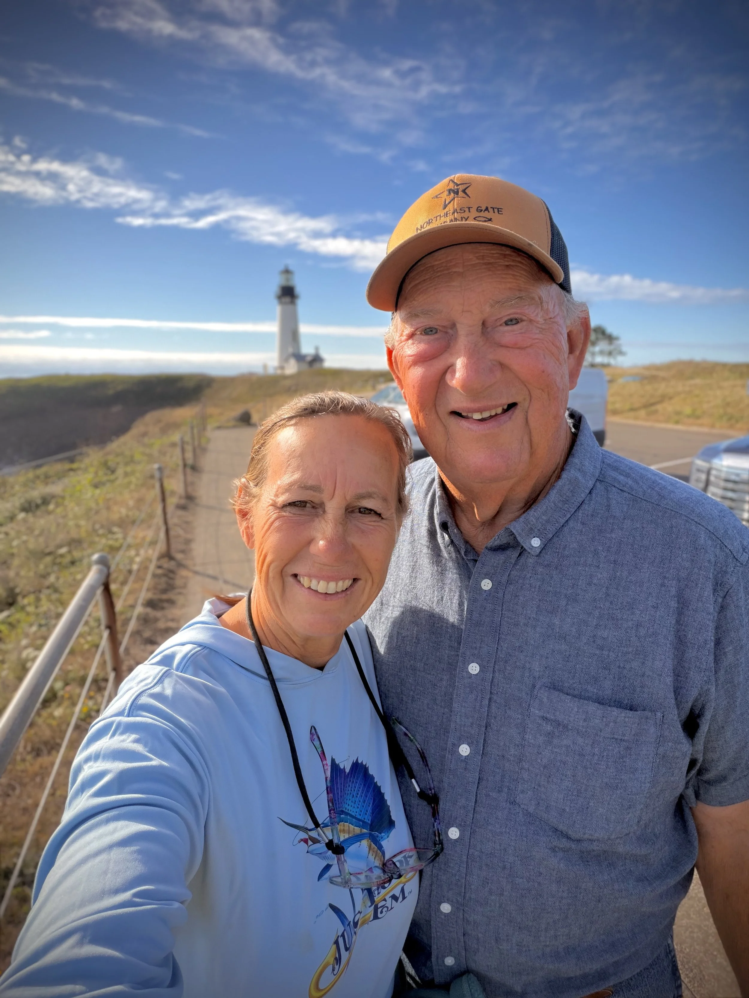 A man and woman taking a selfie outdoors near a lighthouse with a path, parked cars, and grassy landscape in the background on a sunny day.