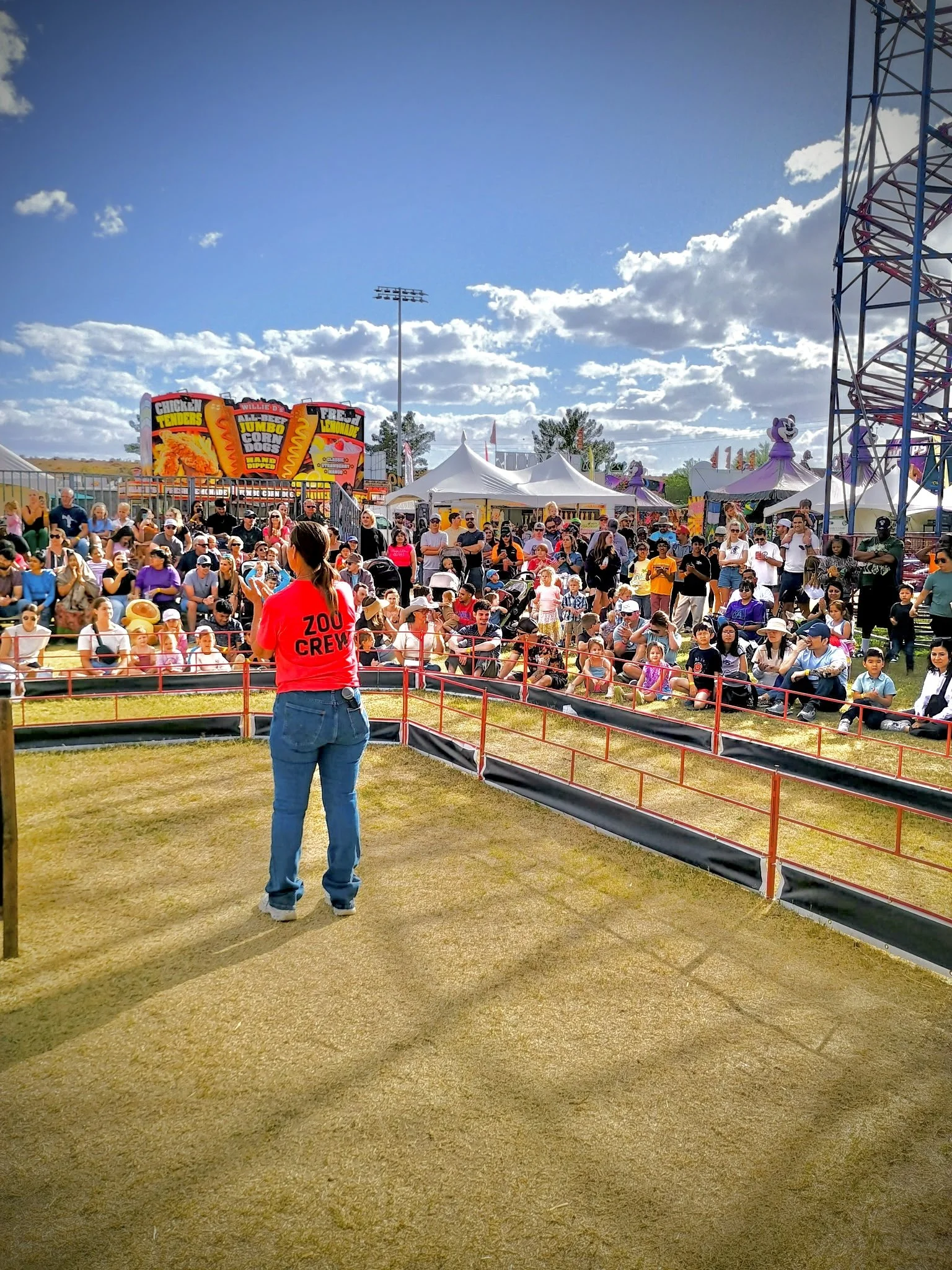 A crowd of people at an outdoor carnival or fair, with a ride and food stalls in the background, and a person in a red shirt labeled 'Zoo Crew' in the foreground.