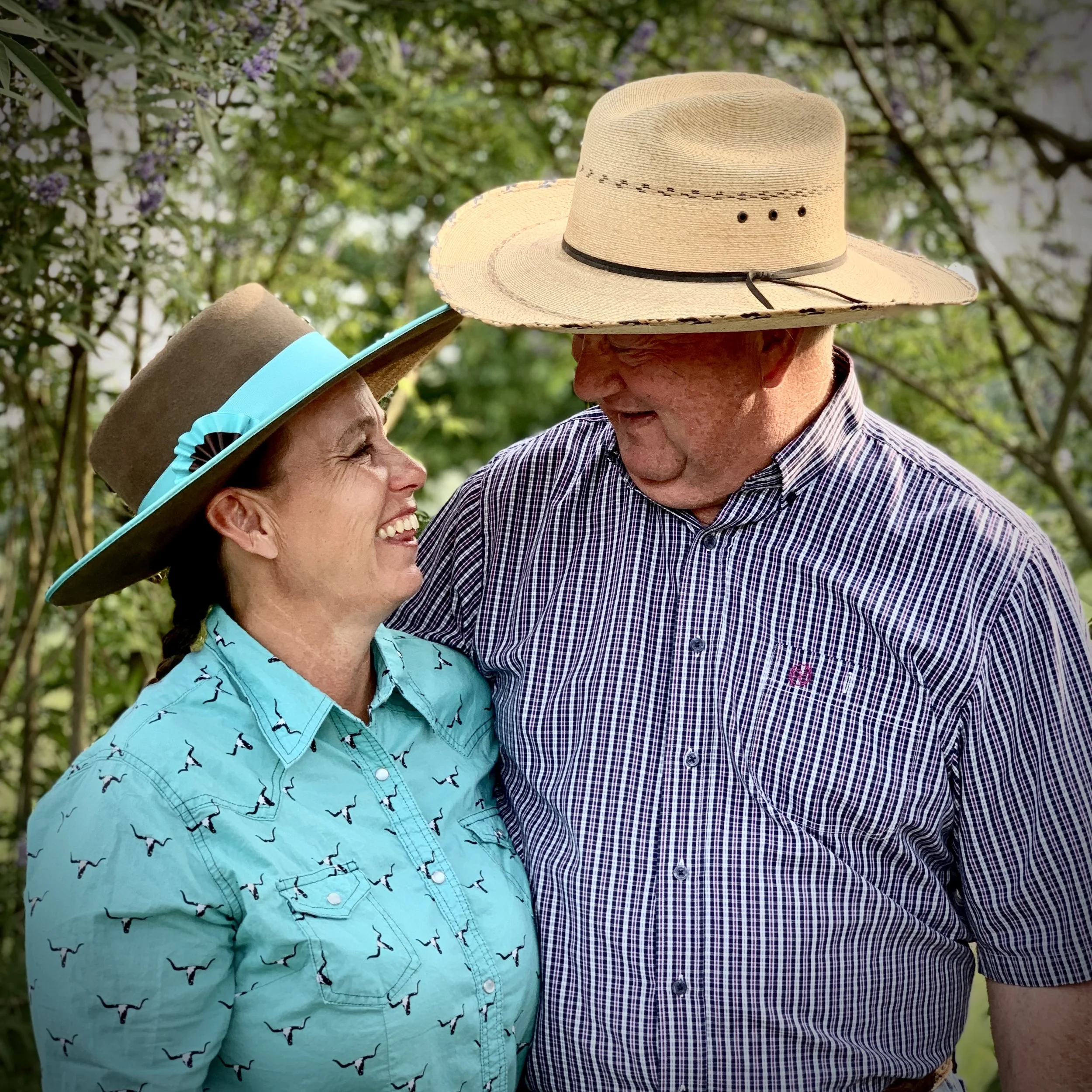 A smiling elderly woman and an elderly man facing each other in a garden, both wearing hats, with the woman in a turquoise shirt and the man in a checkered shirt.