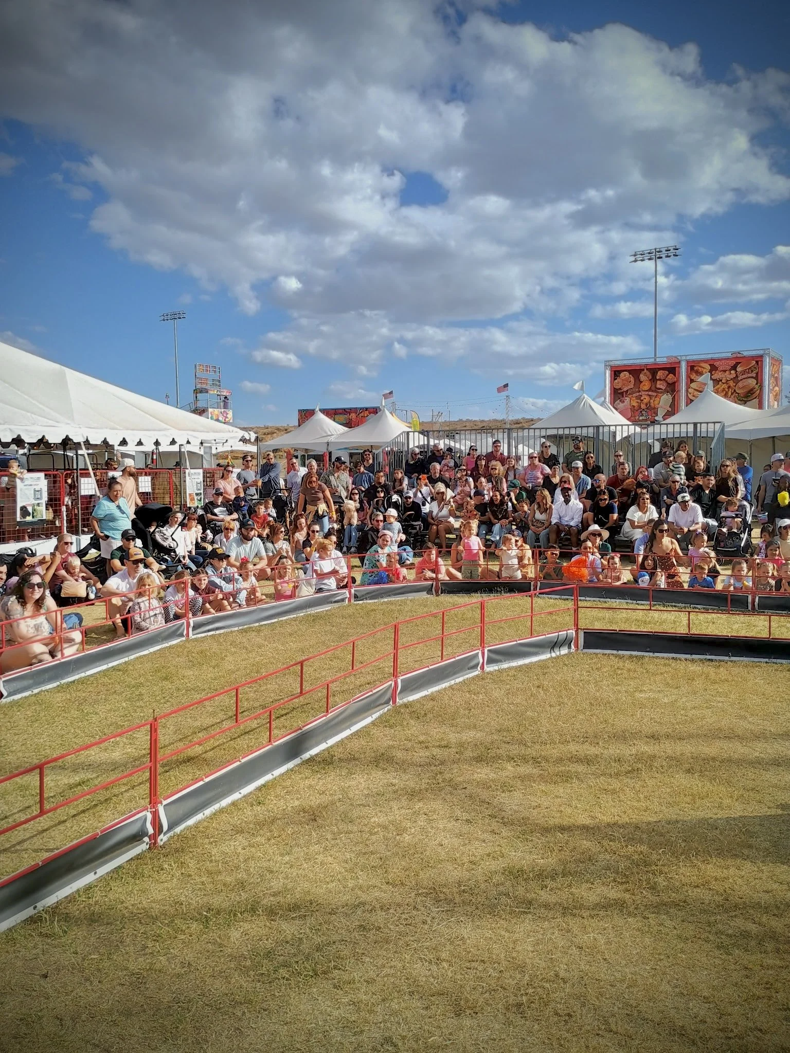 Crowd of people sitting on bleachers at an outdoor event with white tents and large signs depicting food items, under a partly cloudy sky.