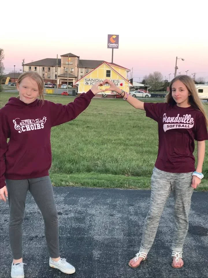 Two girls standing on a parking lot, raising their hands to form a heart shape with the pizza house and Comfort Inn sign in the background during sunset.