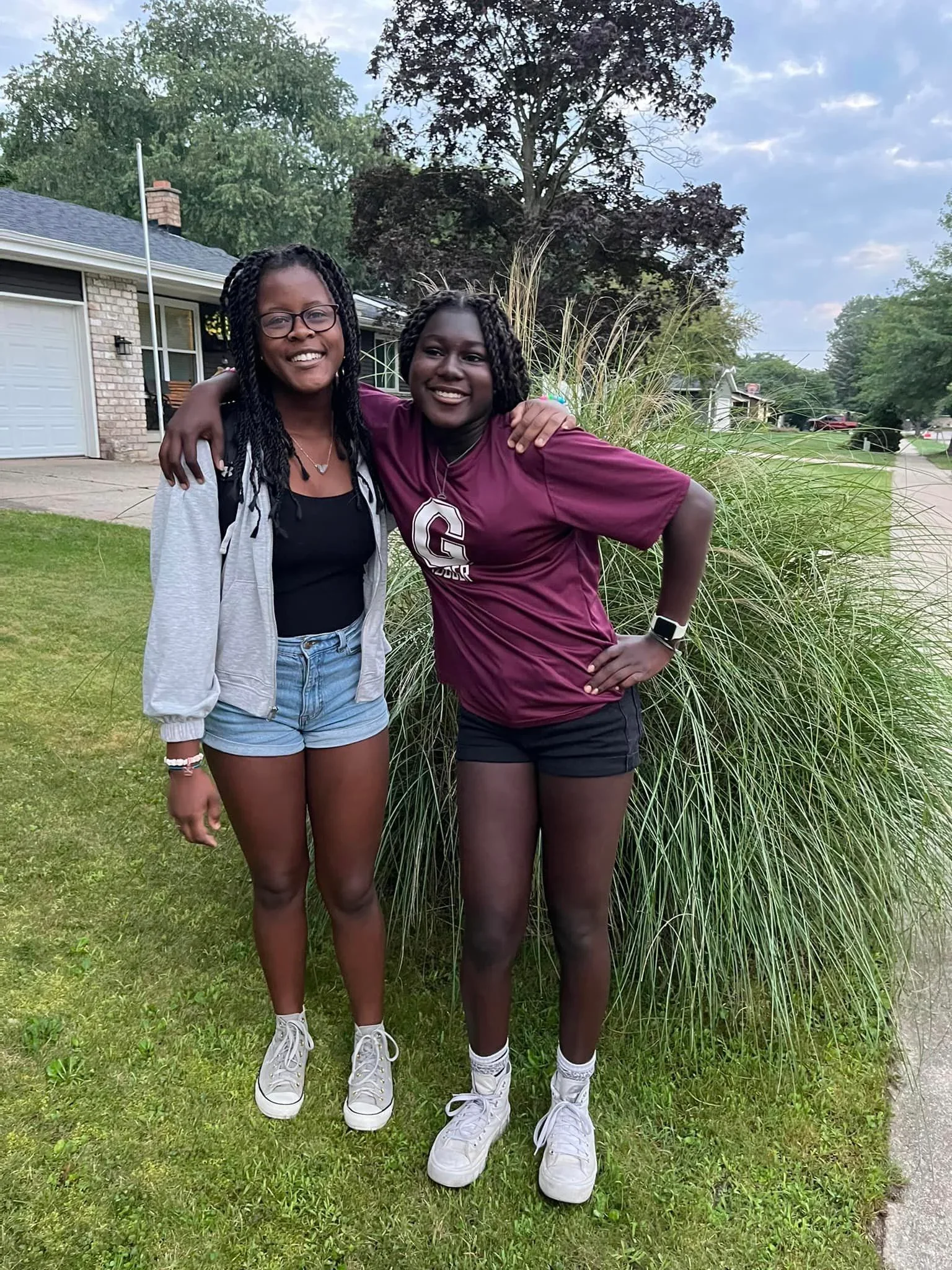 Two young women with dark skin and braided hair standing outdoors in front of a grassy area and tall ornamental grasses, smiling and hugging each other, wearing casual clothes and white sneakers in a residential neighborhood with houses and trees in the background.