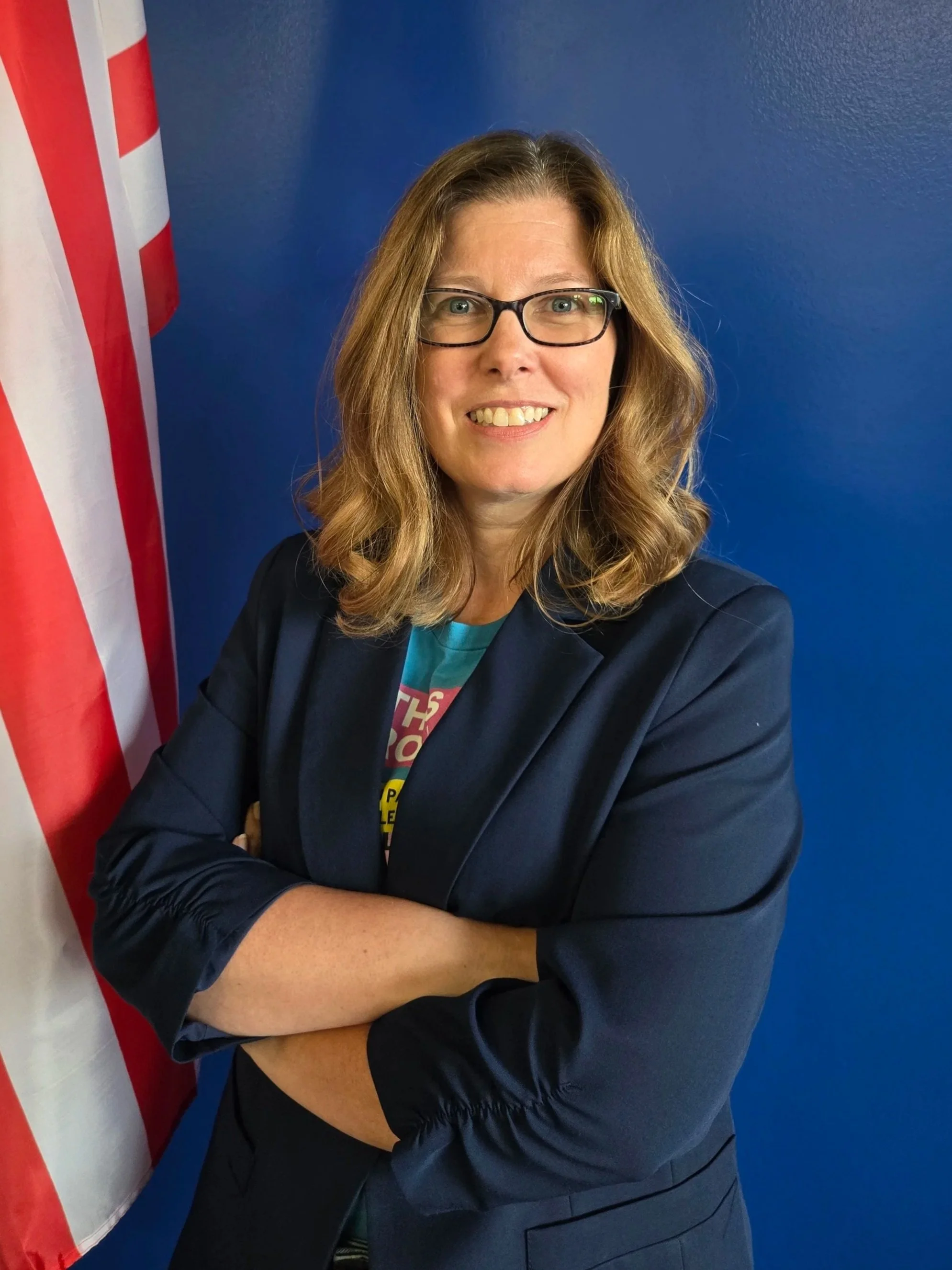 A woman with wavy, shoulder-length hair and glasses smiling while posing with her arms crossed. She is wearing a dark blazer over a colorful shirt. The background features a blue wall and part of a red and white striped flag.