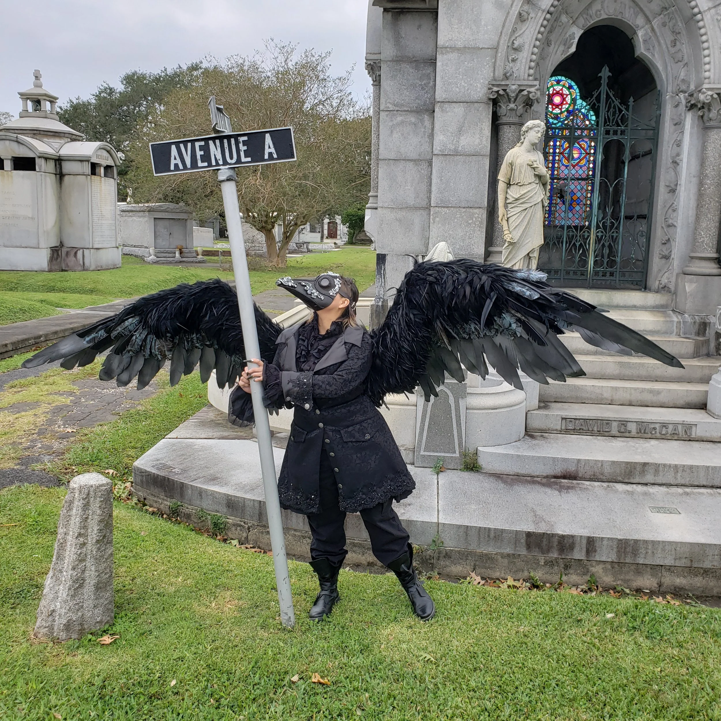 A person dressed in black clothing with large black feathered wings holding a 'Avenue A' street sign in a cemetery with various gravestones, a mausoleum, and a statue of a woman in the background.