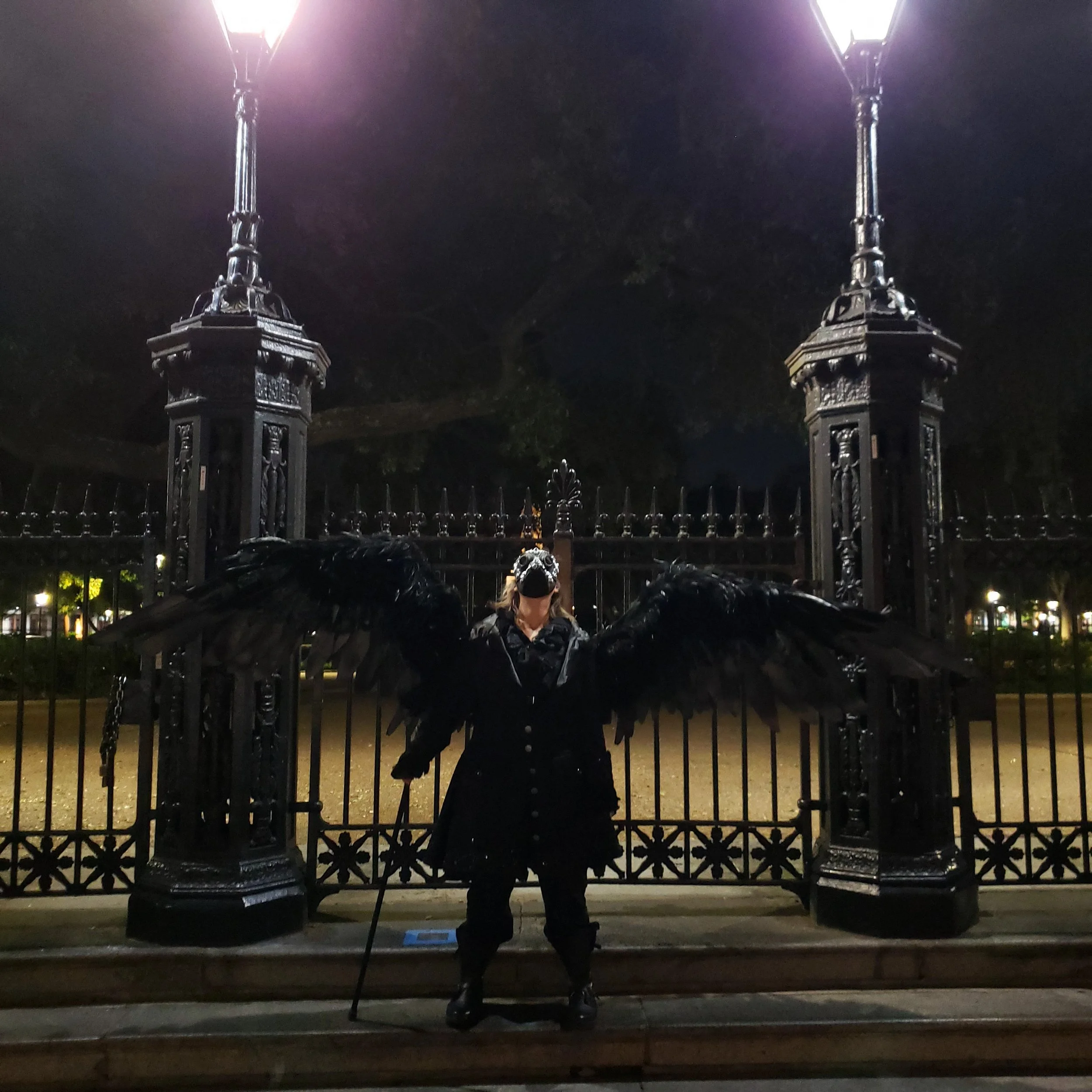 A person with wings standing in front of a black iron gate at night. The person is wearing a black jacket and a hat, holding a walking stick. Two street lamps illuminate the scene from above.