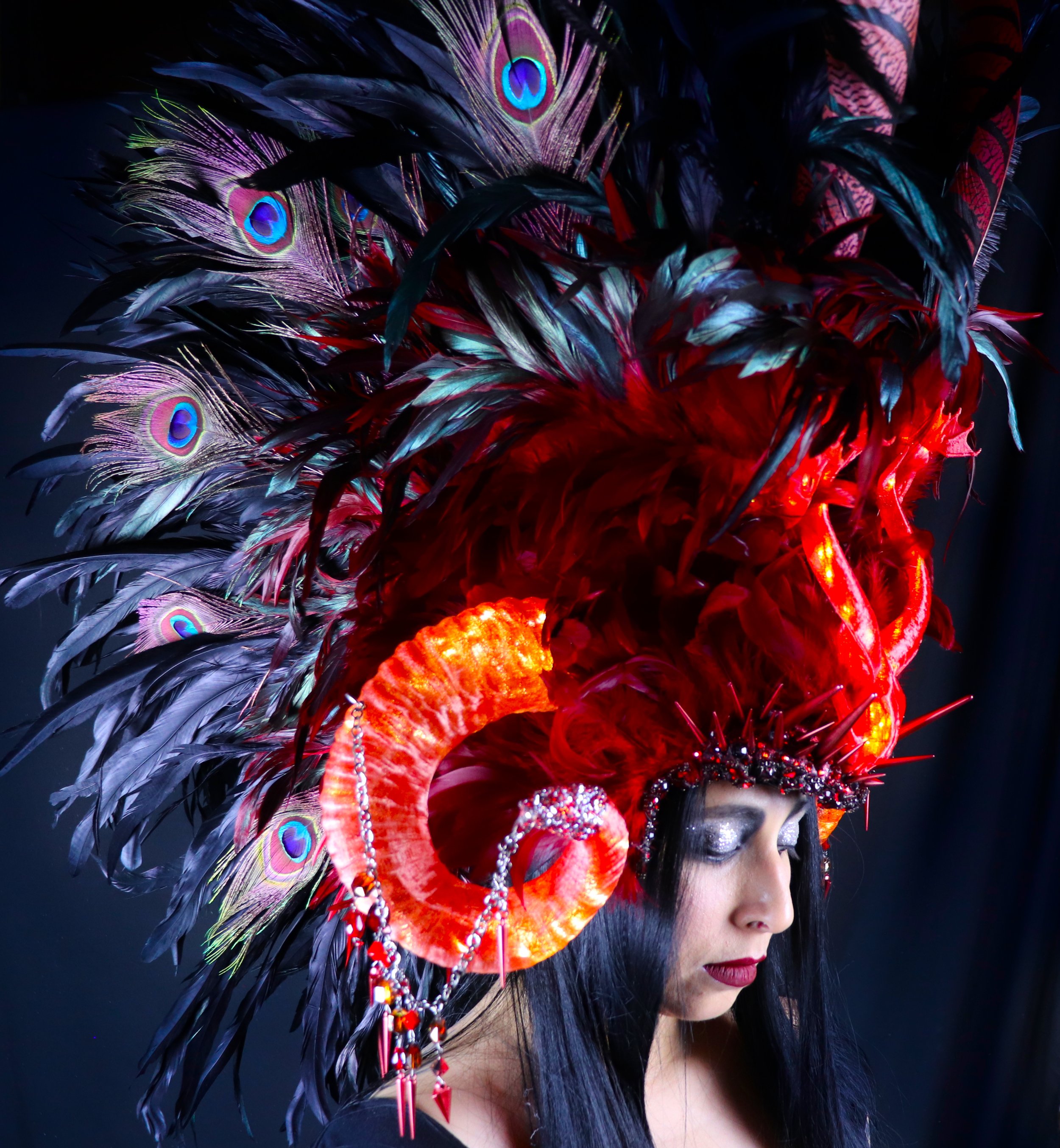 Side profile of a woman wearing a colorful feathered headdress with peacock feather accents, adorned with beads and jewelry, against a dark background.