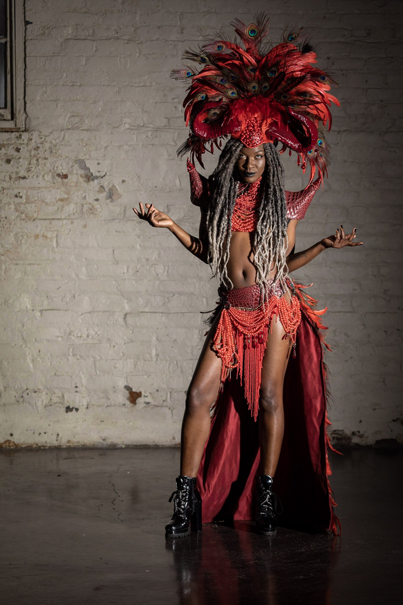 A person with dreadlocks is wearing a vibrant red and black costume with peacock feathers and intricate beadwork, standing against a plain wall with a distressed finish.