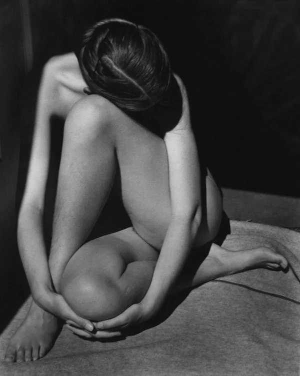A black and white photo of a nude woman sitting on the floor with her legs crossed, holding her ankles with her hands, and her head bent down with her face hidden by her hair.