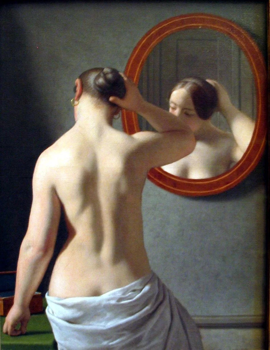 A woman with dark hair styled in two buns looks at her reflection in a round mirror, with her back to the camera, touching the back of her head.