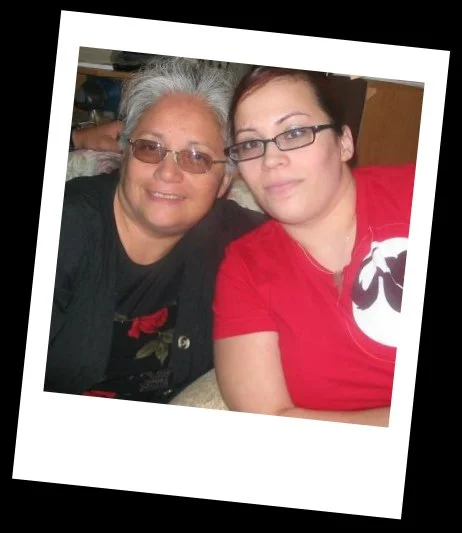 Two women sitting close together, smiling for a photo indoors. One has gray hair and glasses, the other has dark hair and glasses, wearing a red shirt.