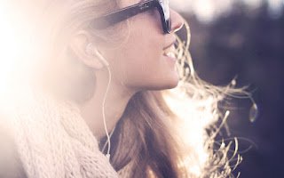 Side profile of a woman with long hair wearing sunglasses and earphones, outdoors with sunlight.