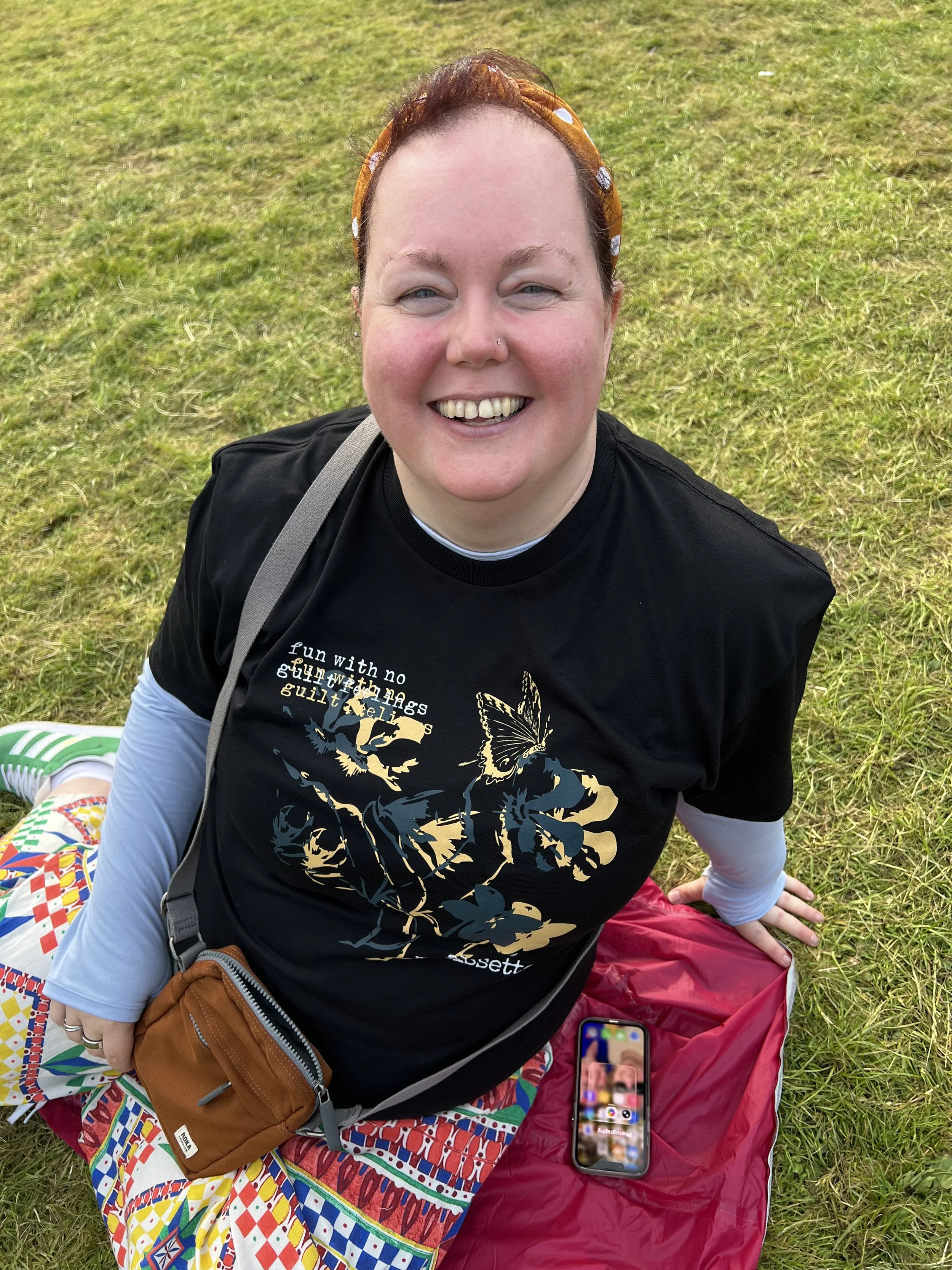 A smiling woman with a nose piercing, wearing a headband and black t-shirt, sitting on colorful patterned fabric on grass, with a smartphone placed nearby and a small brown bag in her lap.