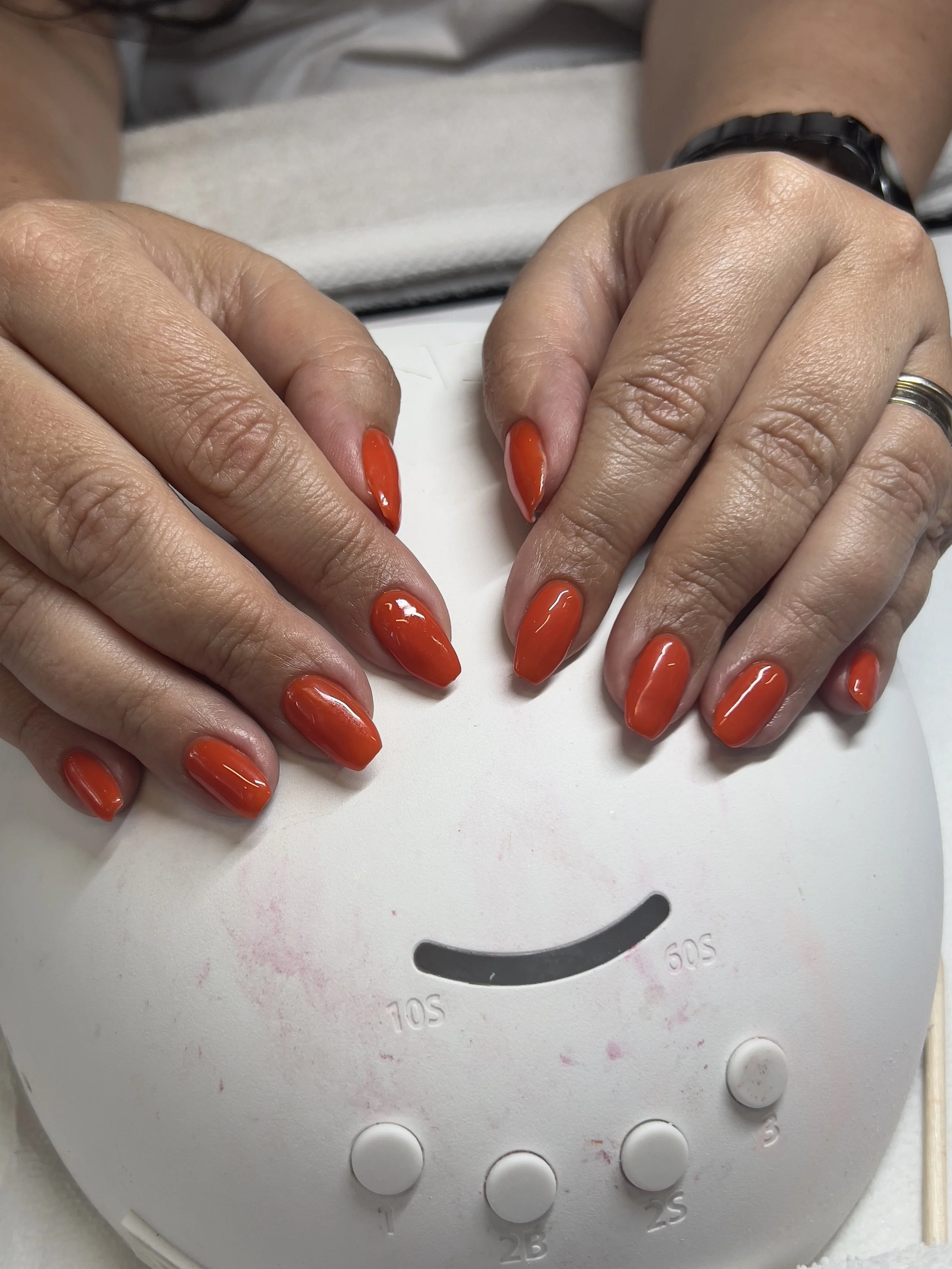 Hands with orange nail polish resting on a white surface, with a partial view of a white device with buttons in the background.