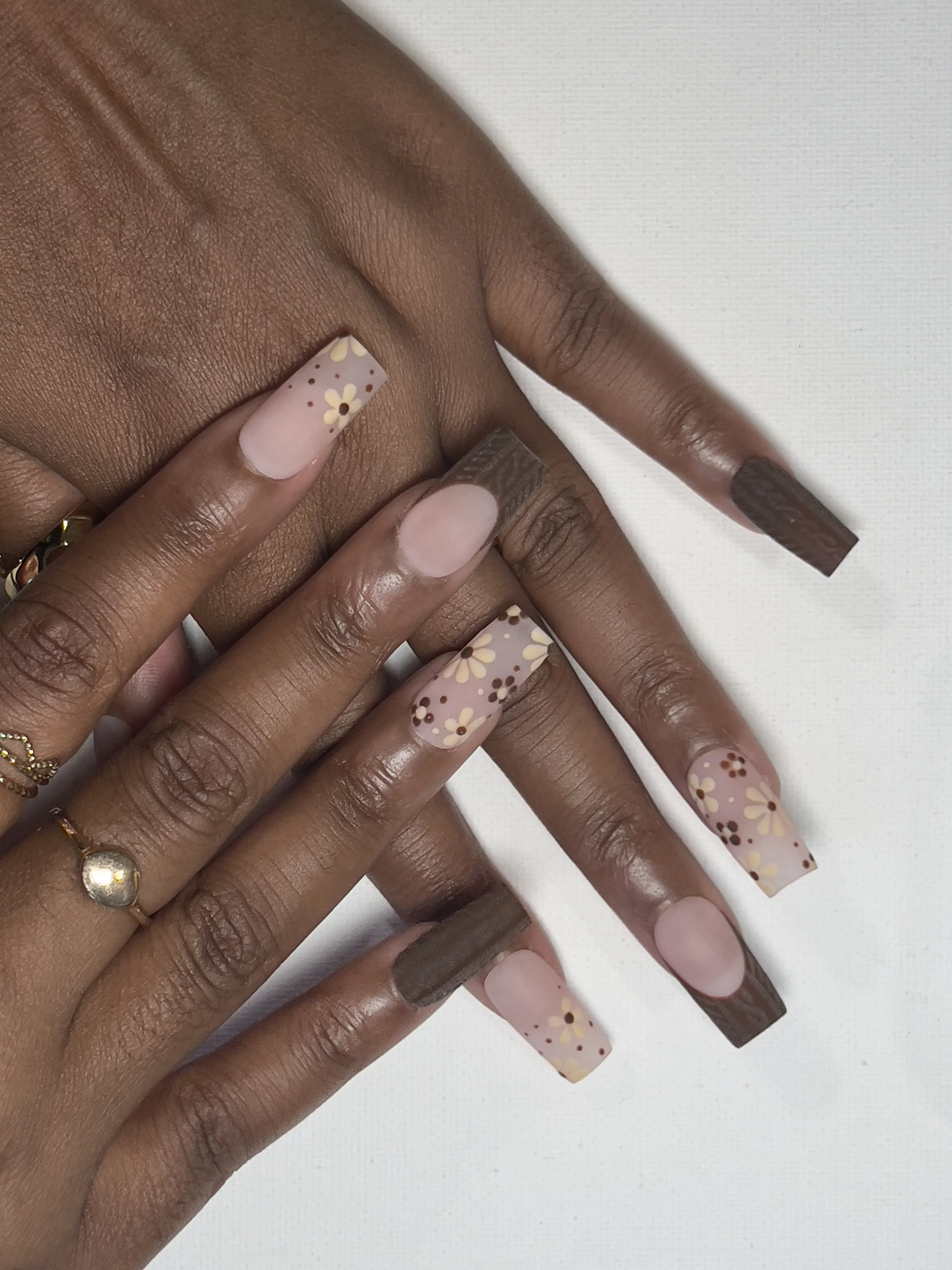 Close-up of hands with dark skin, showcasing manicured nails with floral designs and a matte brown color on some nails, against a white background.