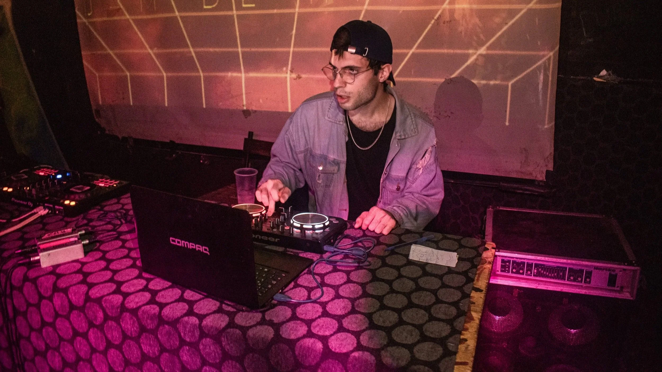 Joven DJ mezclando música en una cabina, usando audífonos, con un fondo de pantalla de proyección en la pared, en un ambiente de fiesta o club nocturno, con una superficie de mesa con patrones y equipo de sonido.