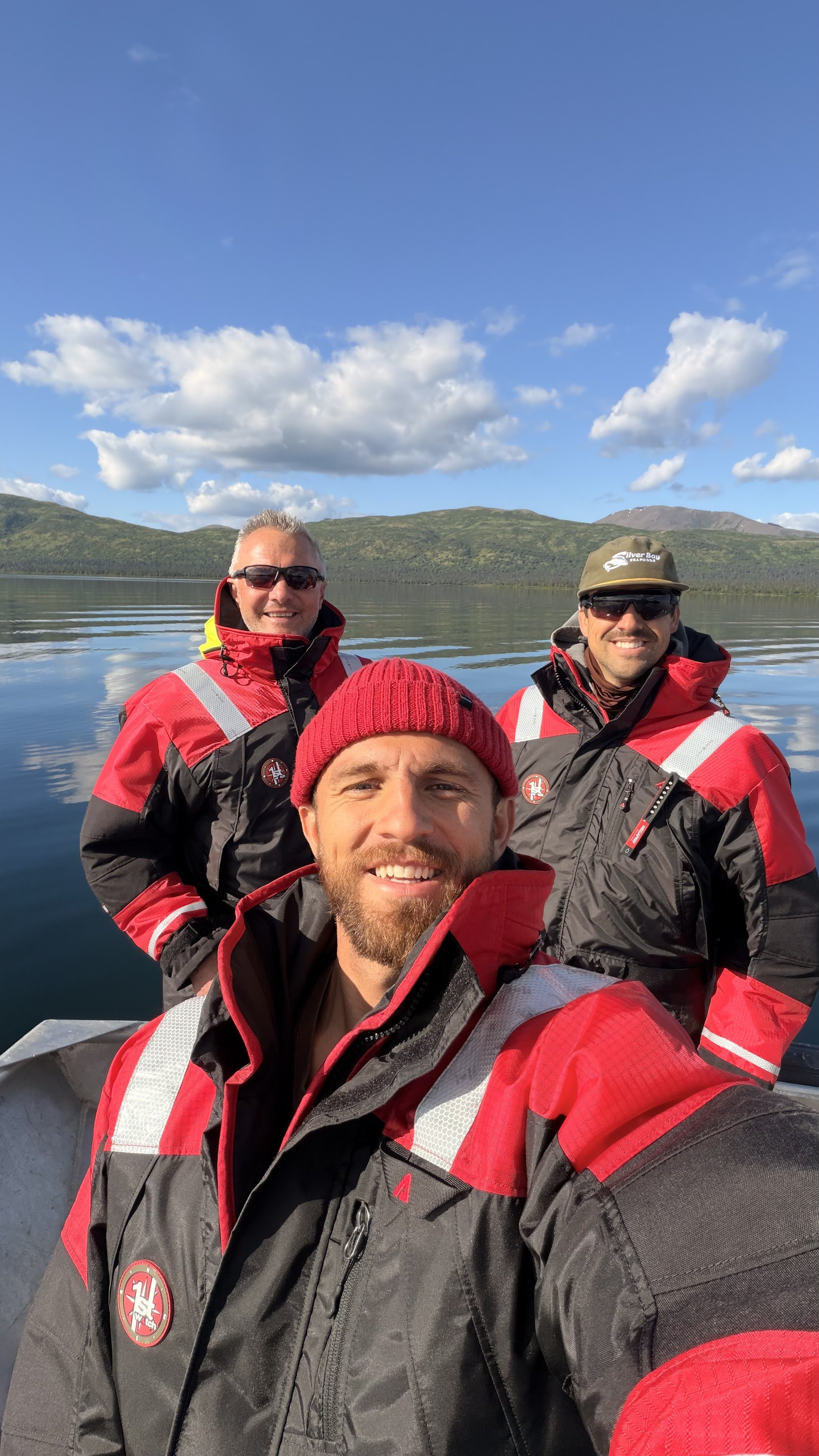 Three men in red and black waterproof jackets taking a selfie on a boat with a lake and mountainous landscape in the background, under a blue sky with white clouds.