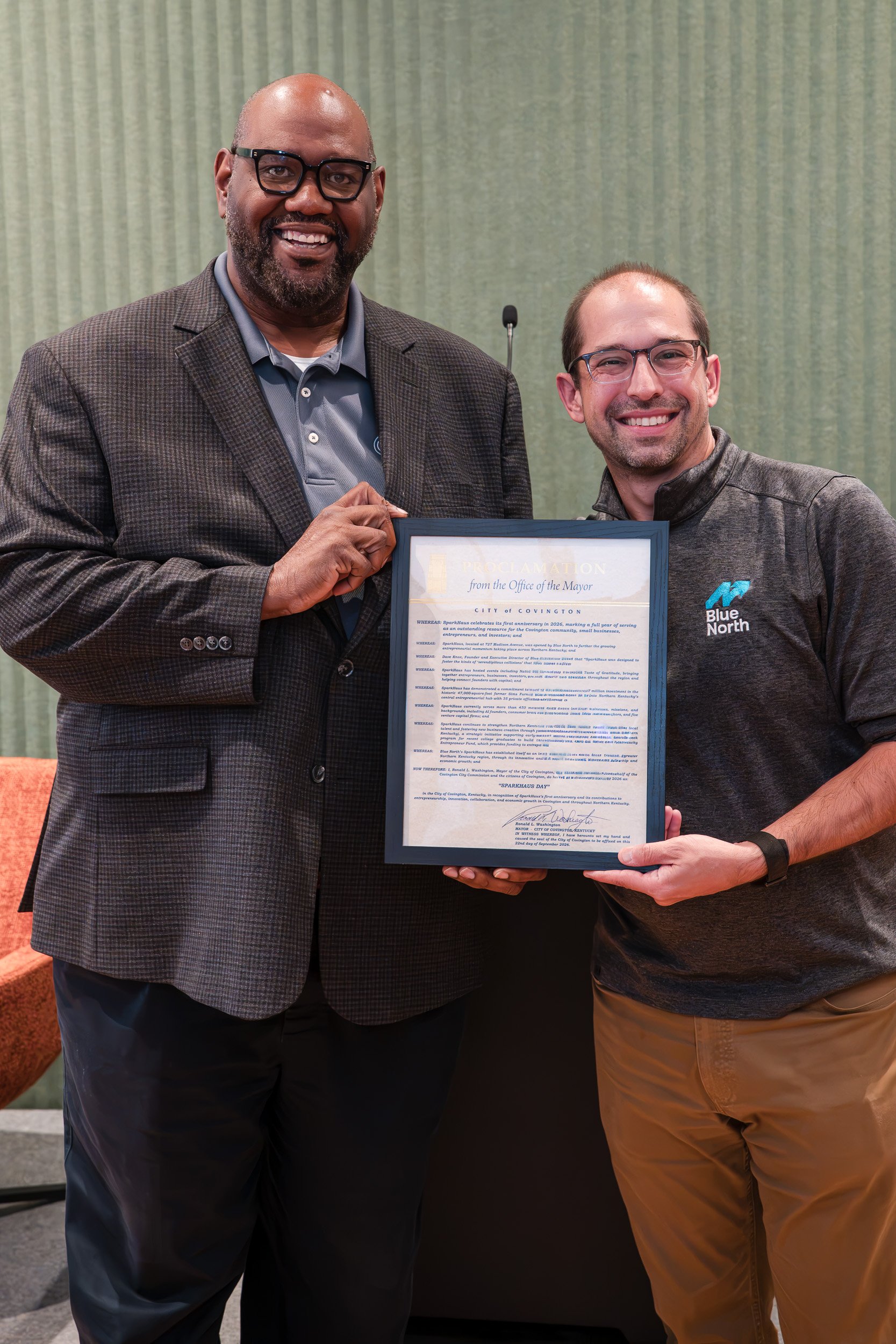 Two men smiling and holding a framed proclamation at an indoor event, with one wearing glasses, a gray blazer, and a gray shirt, and the other wearing glasses, a dark gray jacket with a blue logo, and tan pants.