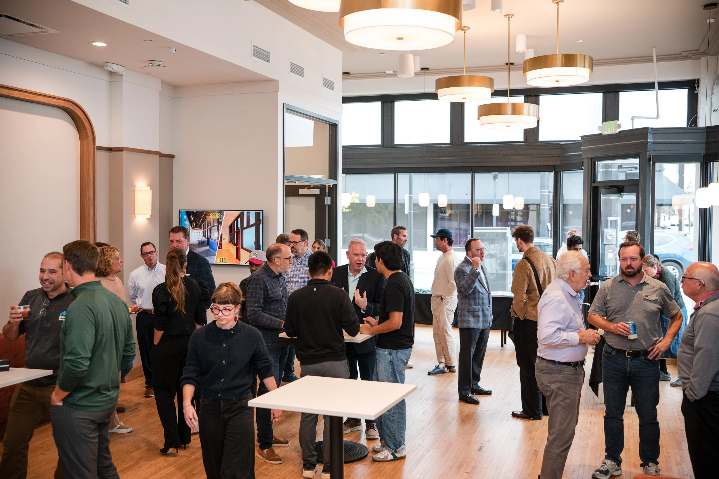 A group of people socializing at an indoor event, with large windows, hanging circular lights, and a television on the wall.