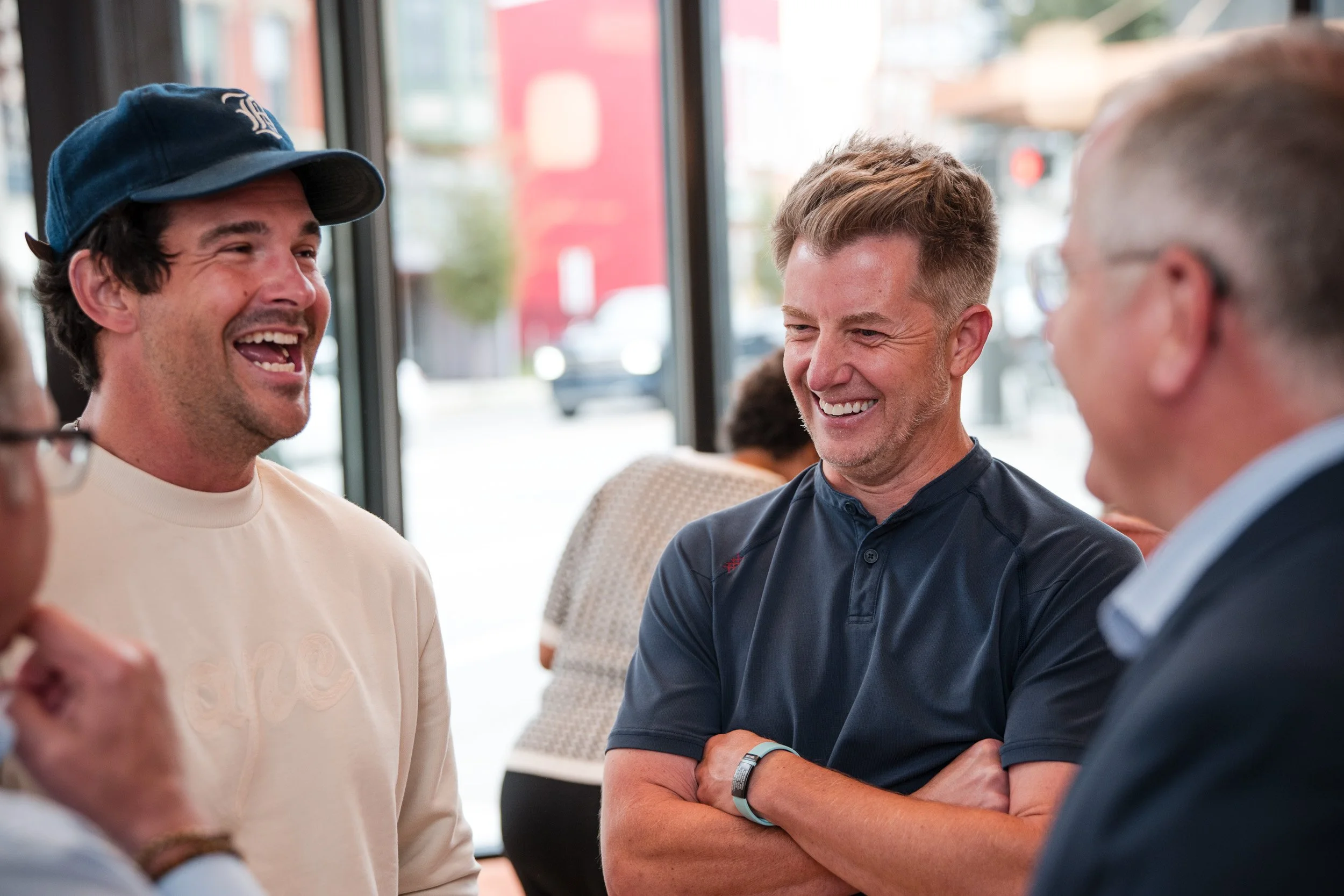Four men are standing together indoors, smiling and engaging in conversation. The man on the left wears a baseball cap and a light-colored shirt. The man in the center is dressed in a dark shirt with his arms crossed. The other individuals are partia